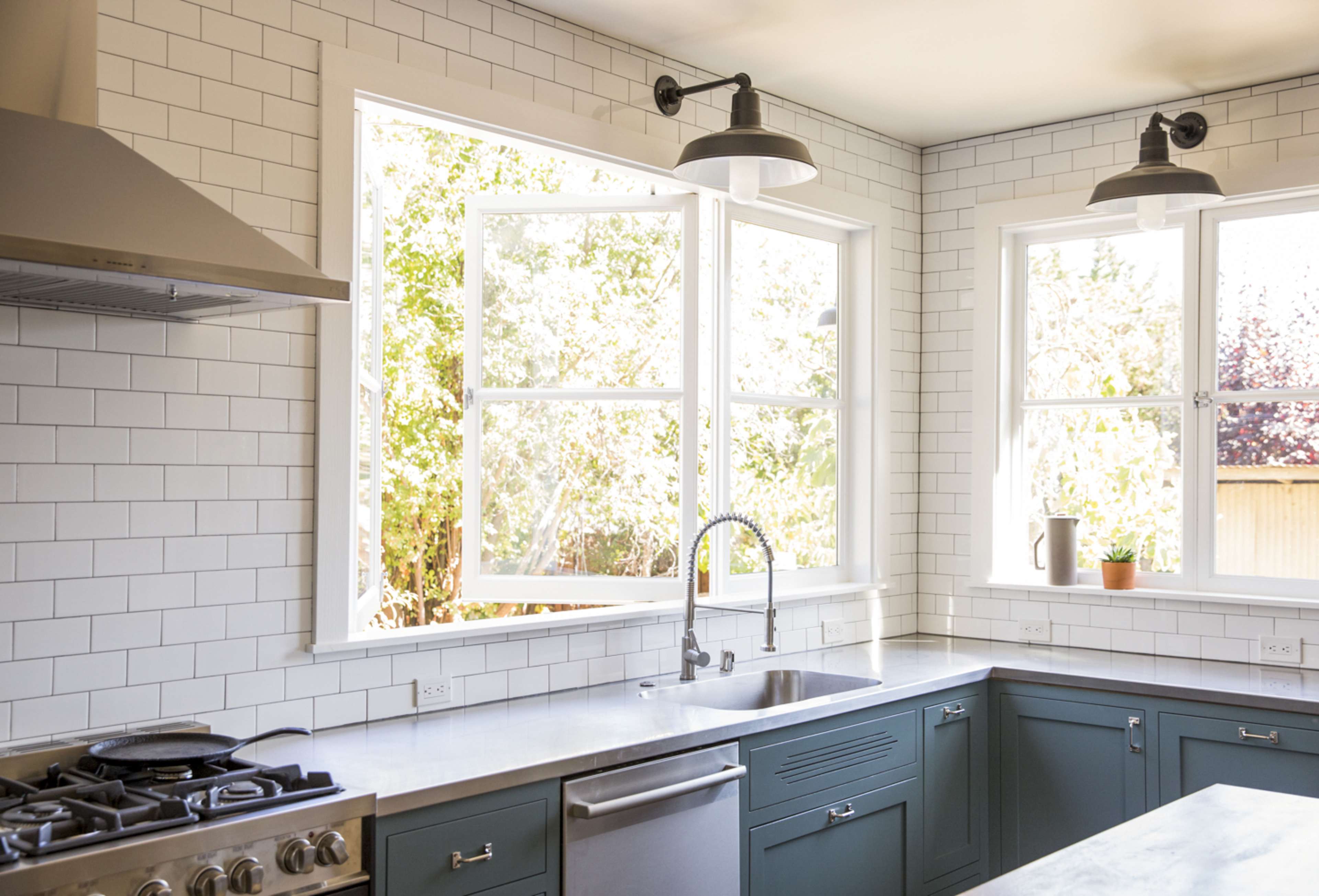 A modern kitchen features teal cabinets, a stainless steel sink, and large windows allowing natural light to enter.