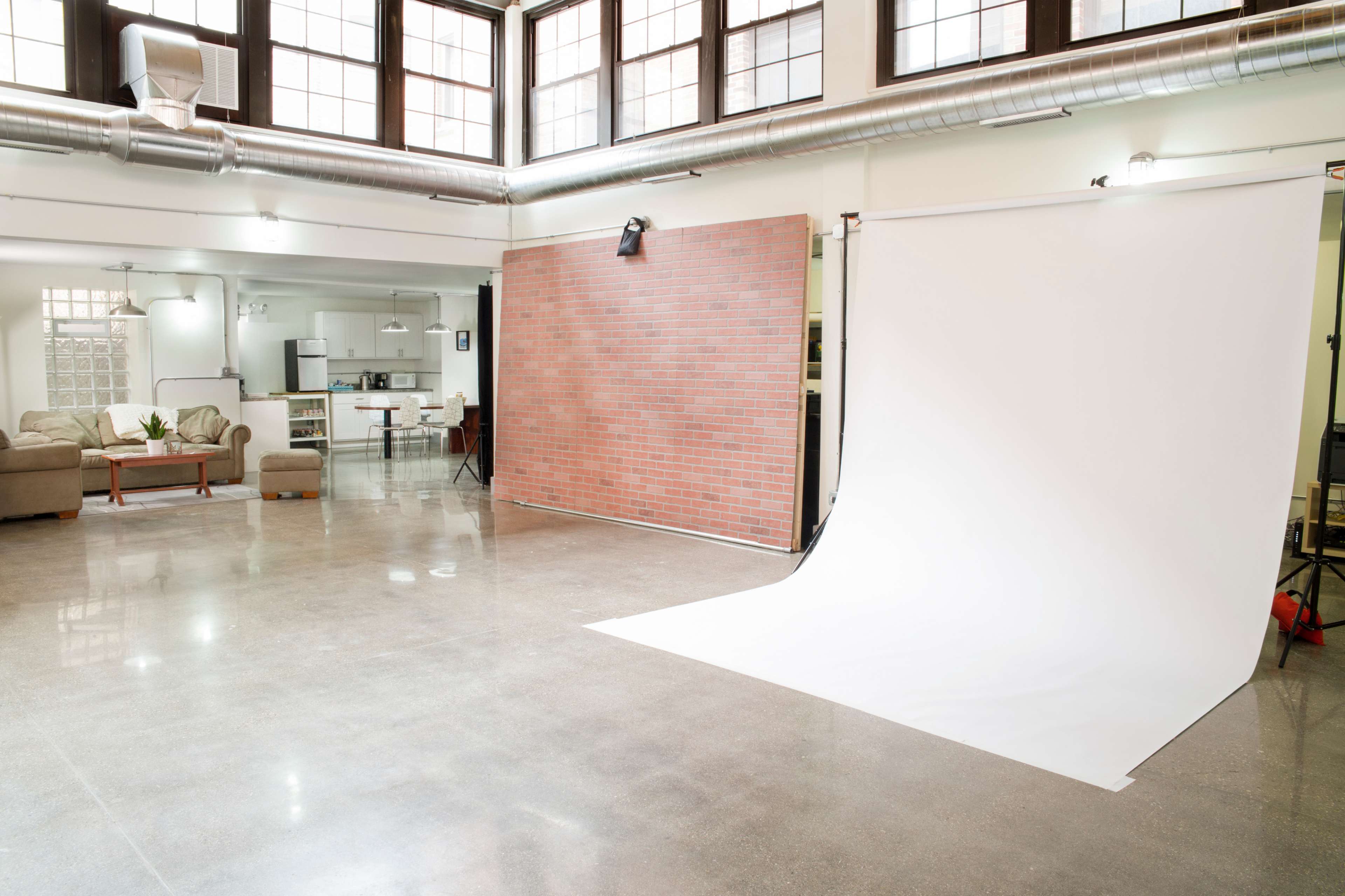 The image shows a spacious studio with a smooth concrete floor, a white backdrop, and a brick wall, along with a seating area and a kitchen visible in the background.