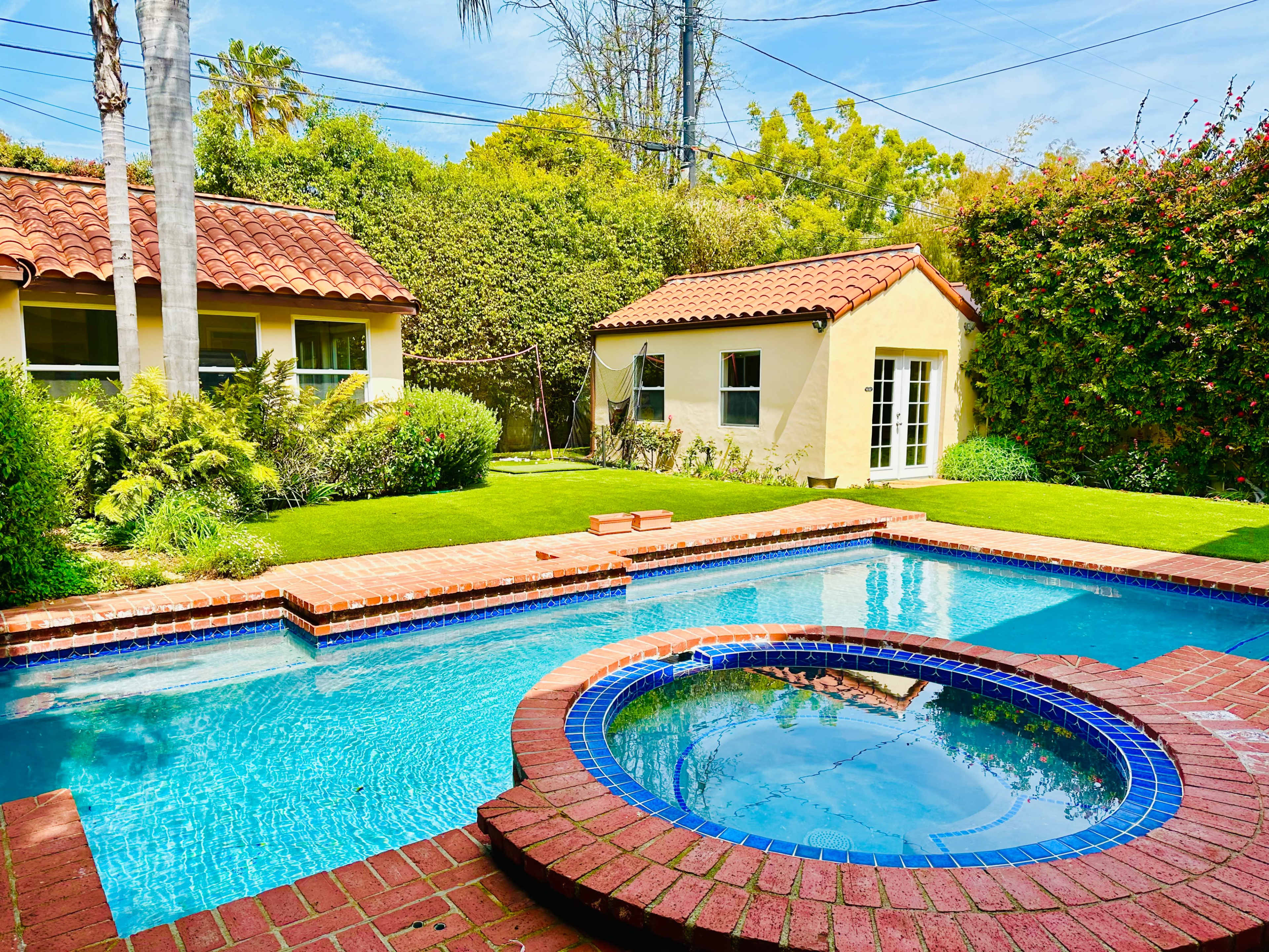 The scene features a backyard with a swimming pool and a jacuzzi, surrounded by green lawns and two adjacent houses.