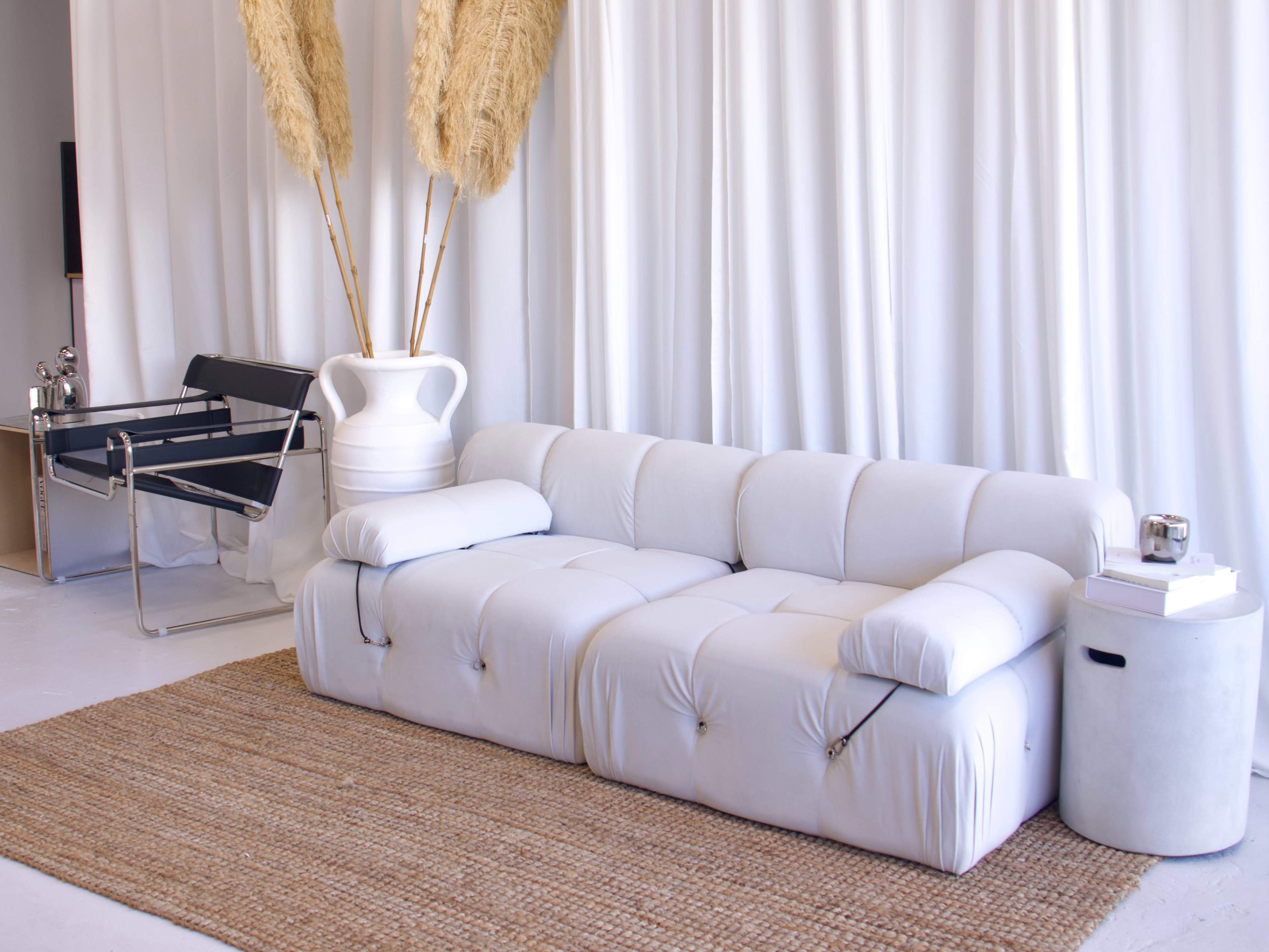 A modern living room features a white sectional sofa, a minimalist side table, and tall pampas grass in a large vase against a backdrop of white curtains.