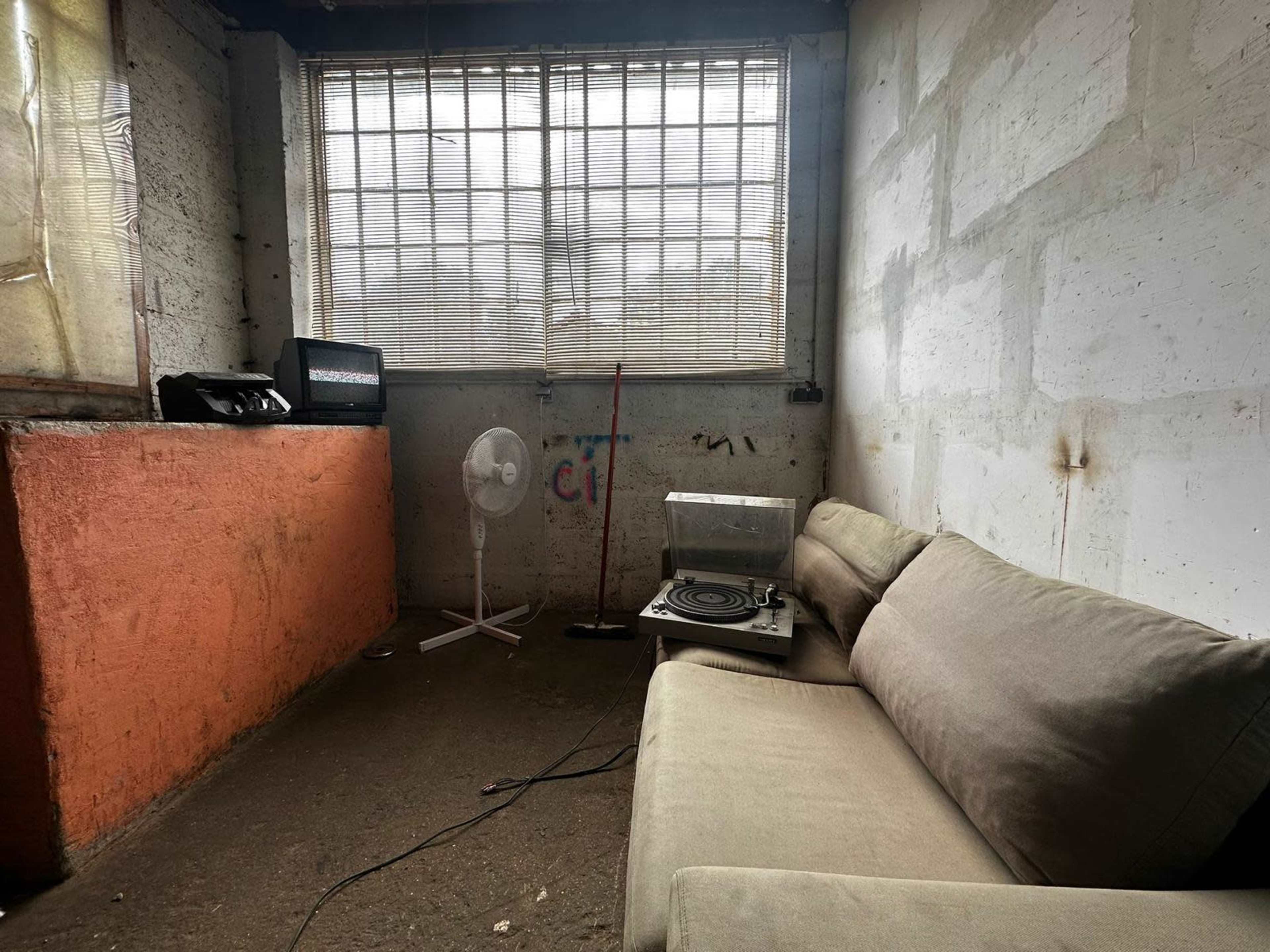 The image shows a sparsely furnished indoor space featuring a light-colored couch, a fan, an old television, and a turntable situated against a bare concrete wall with a window partially covered by a curtain.