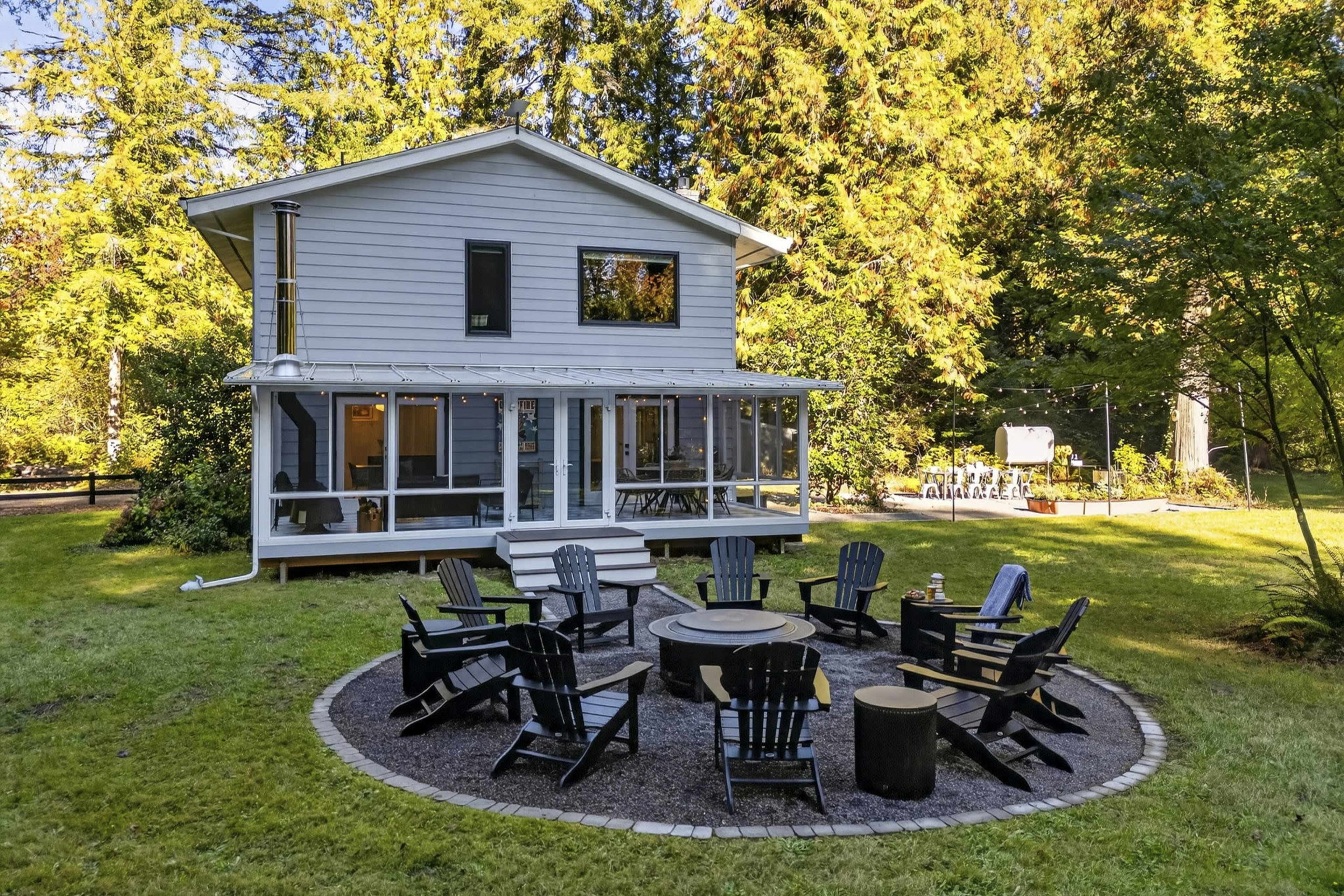 A two-story house with a porch and a circular fire pit surrounded by Adirondack chairs is set in a forested area.