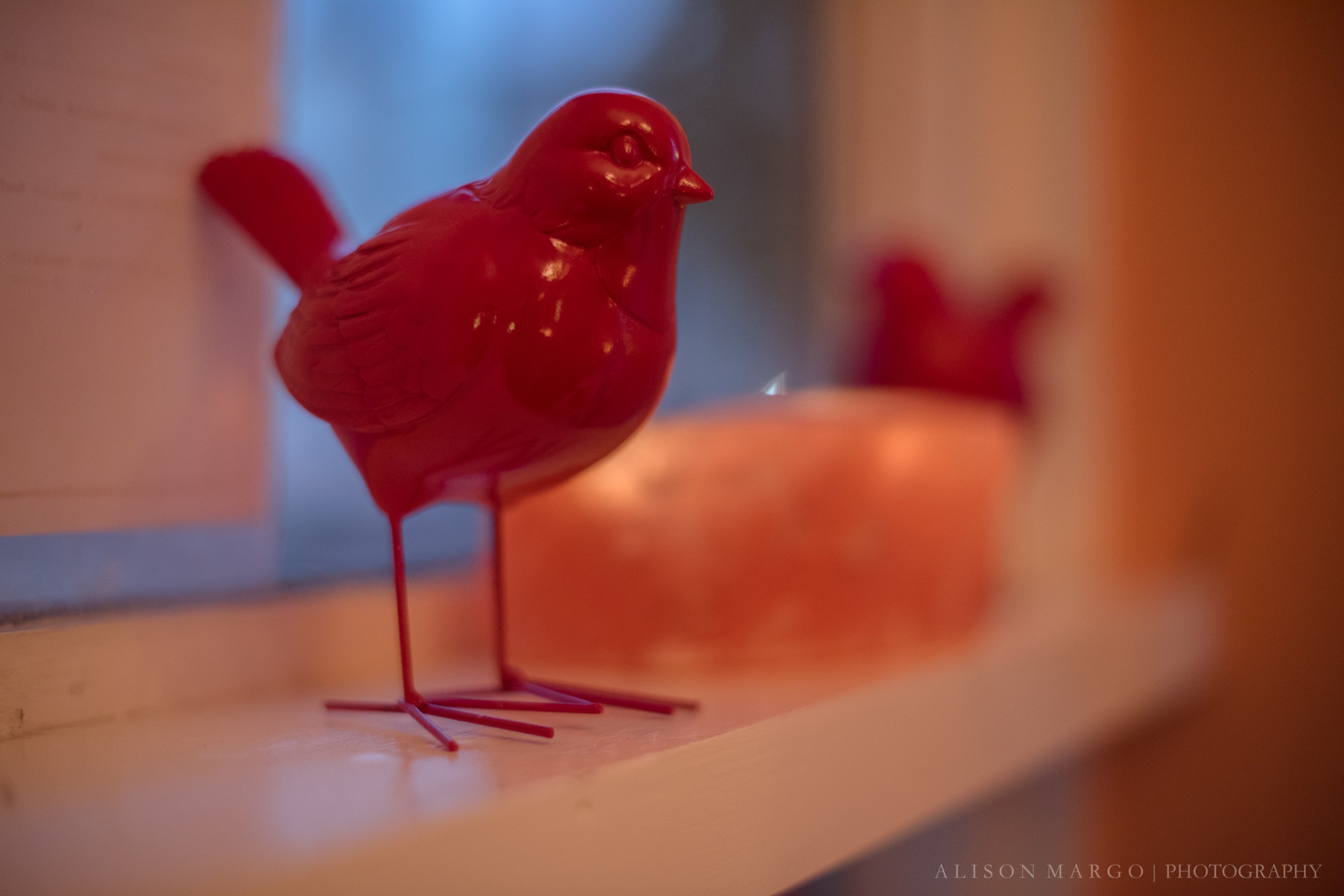 A bright red bird figurine stands on a windowsill, with another figurine blurred in the background.
