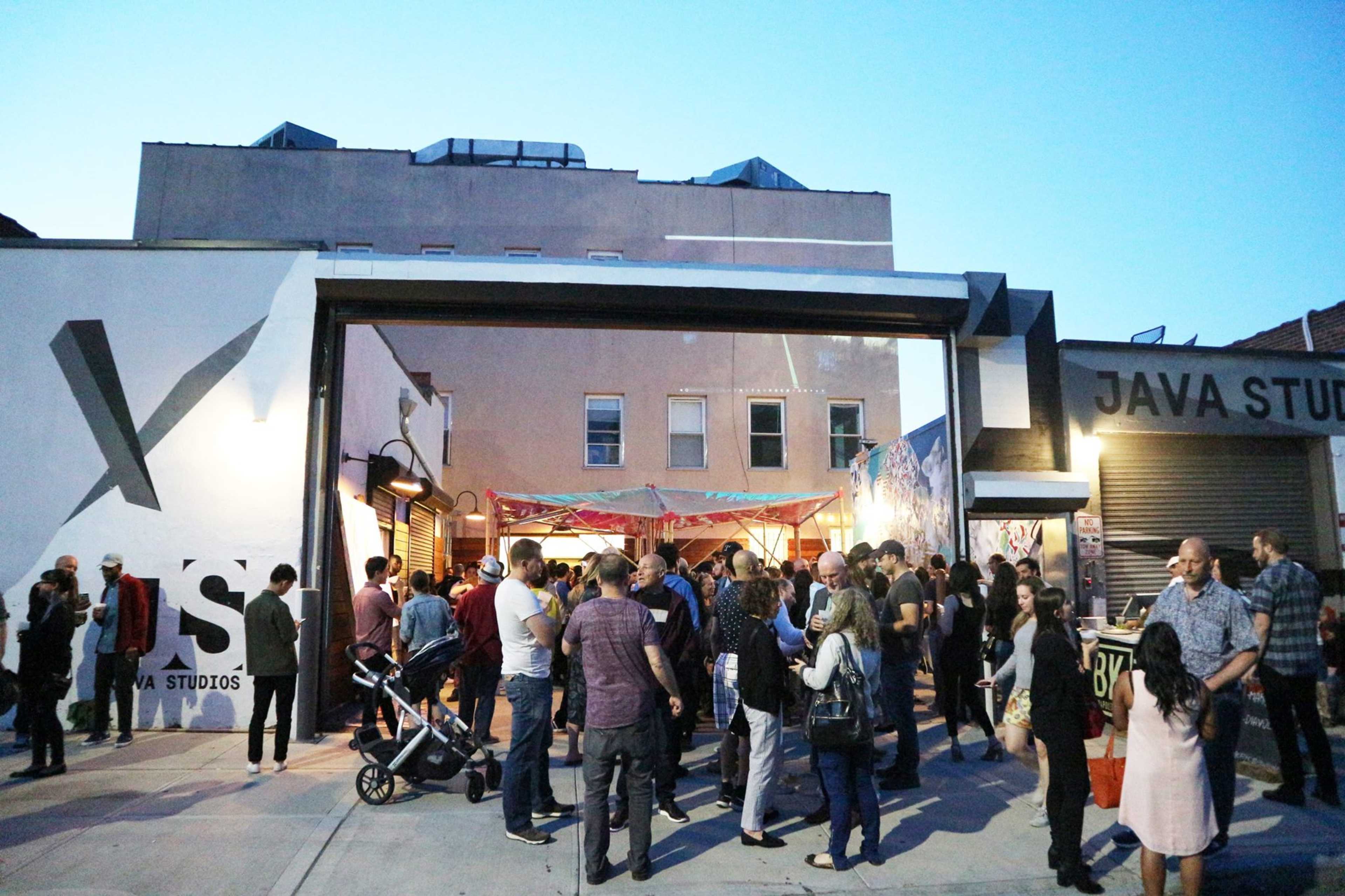 A crowd gathers outside Java Studios, with people mingling and chatting under the evening sky.