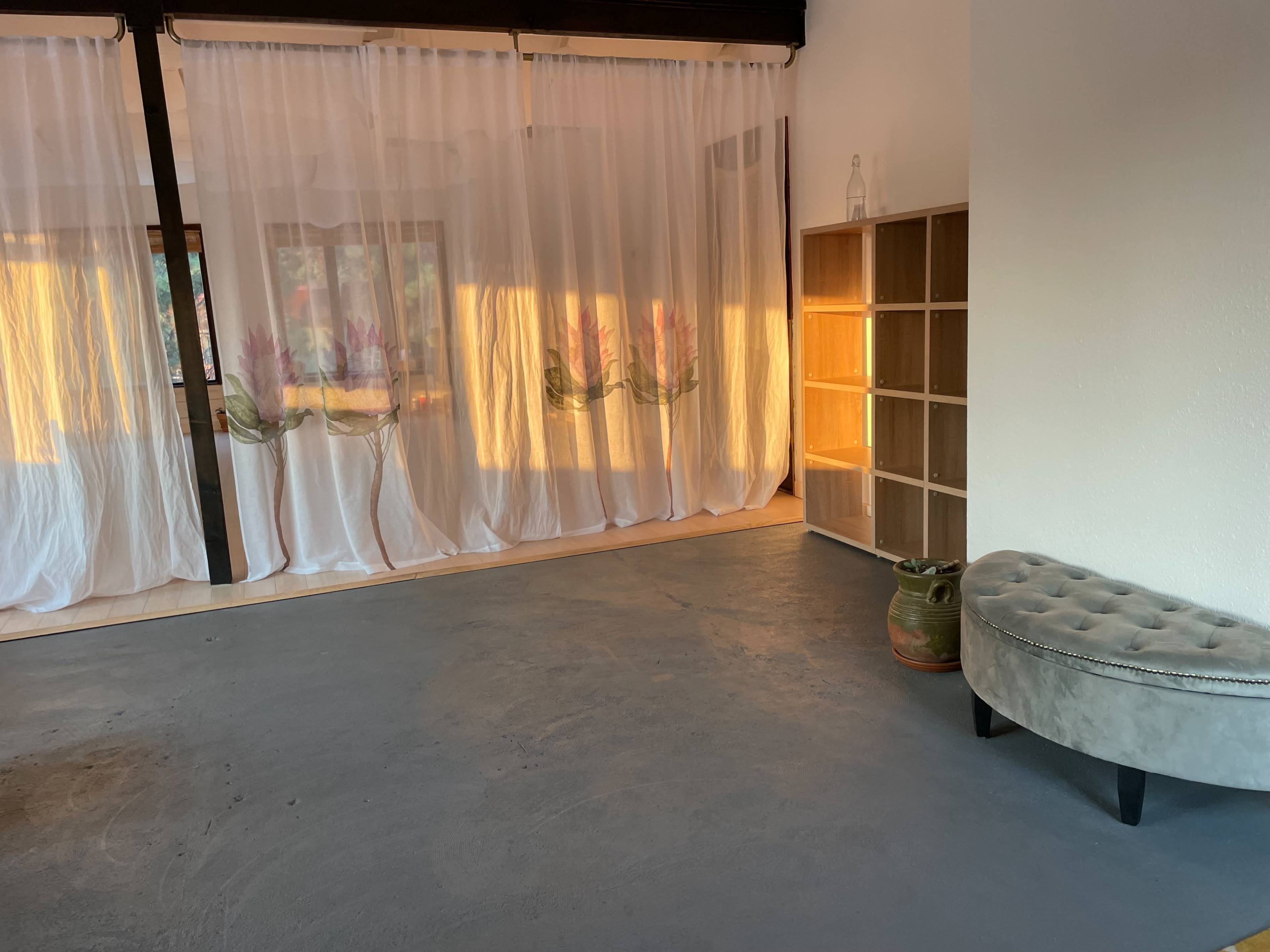 A spacious room with sheer white curtains, a light-colored bookcase, and a tufted bench against a wall, illuminated by warm sunlight.