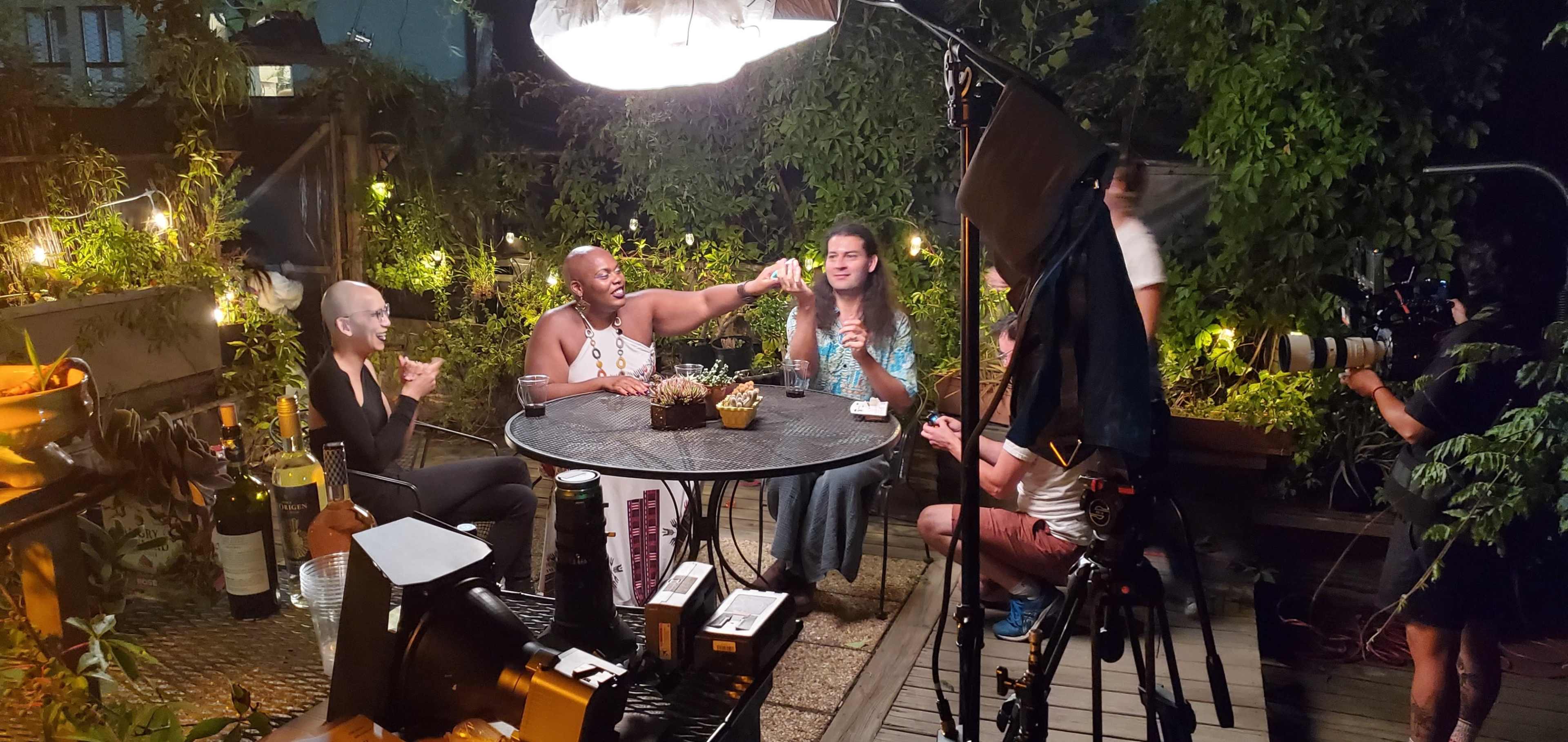 A group of five people gathers around a table in a dimly lit outdoor space adorned with string lights and greenery.