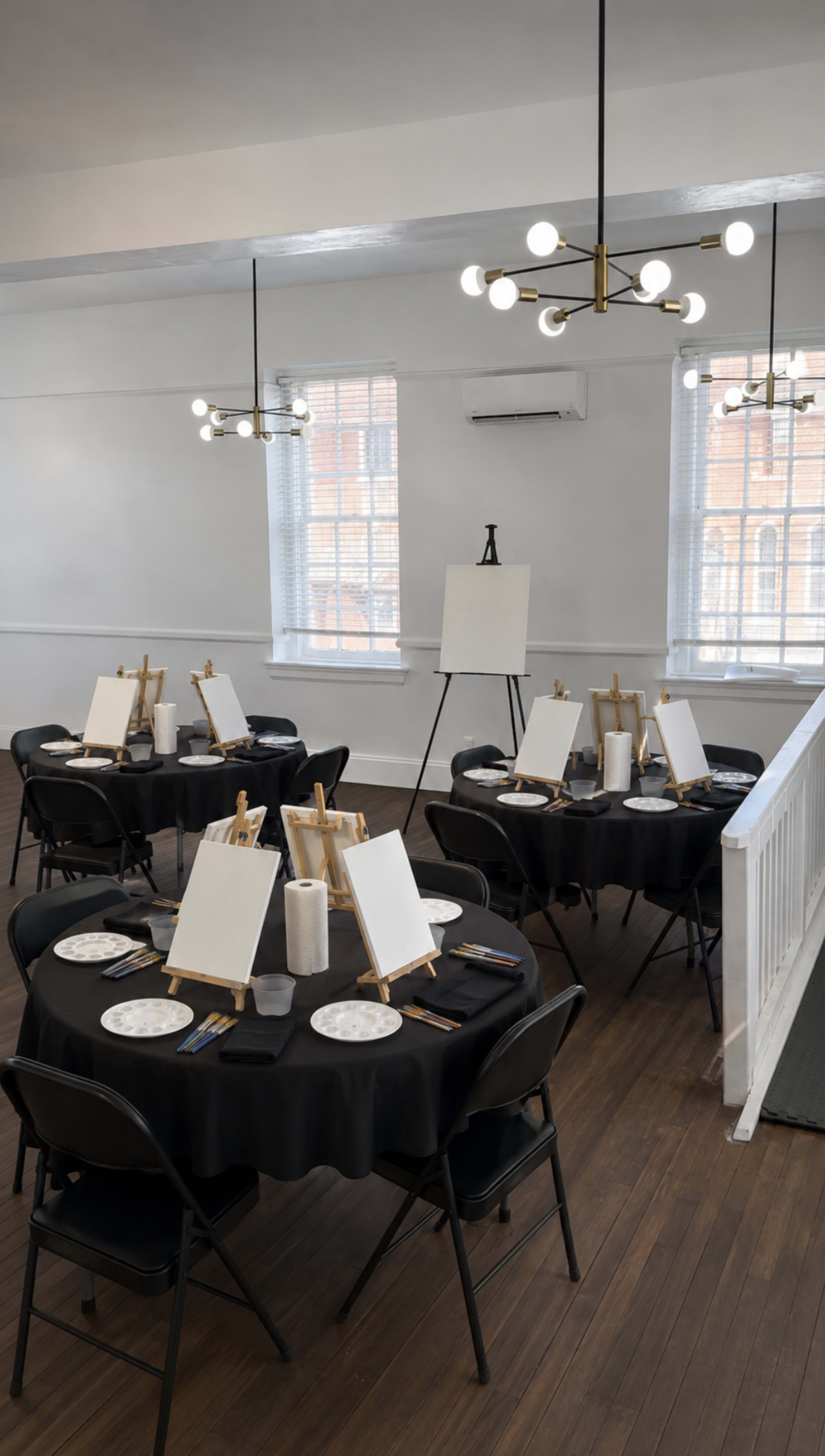 The scene shows a well-lit room set up for an art class, featuring several round tables with blank canvases on easels and plates at each setting.