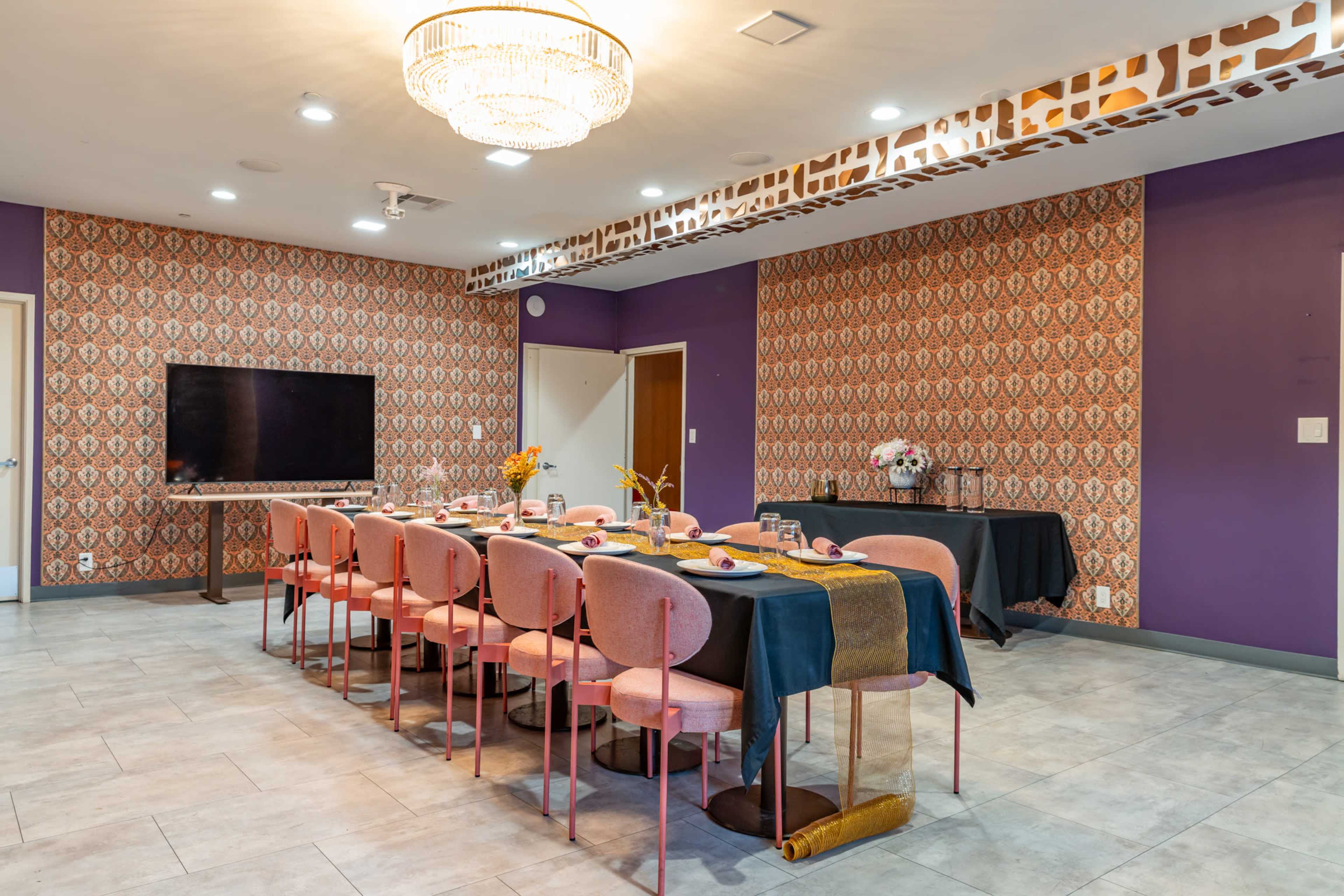 The image shows a modern dining room with a long table set for a meal, surrounded by pink chairs, featuring patterned wallpaper and a large chandelier.
