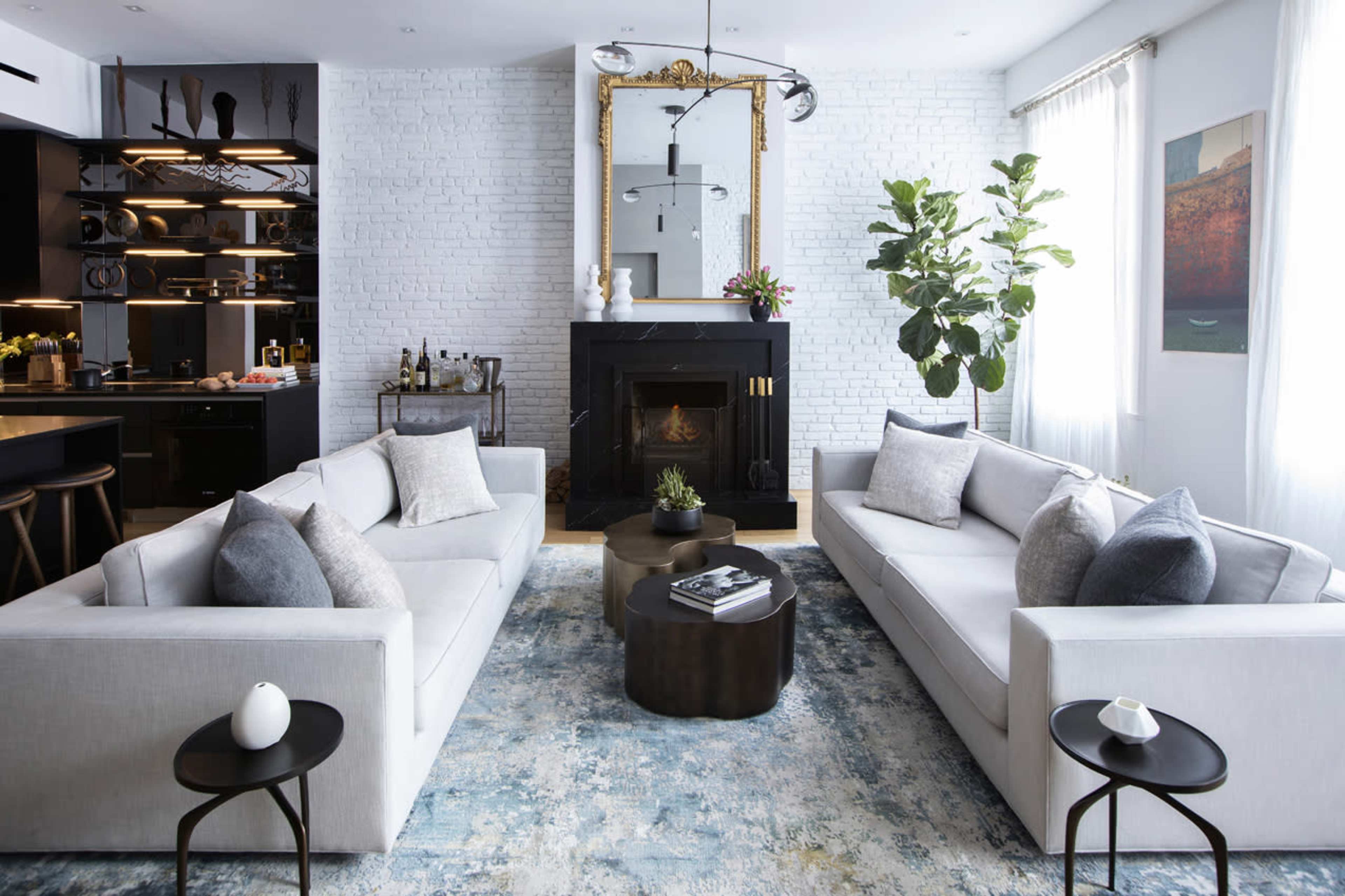 The image shows a modern living room with two sofas facing each other, a round coffee table in the center, and a fireplace with a mirror above it, surrounded by plants and decorative items.