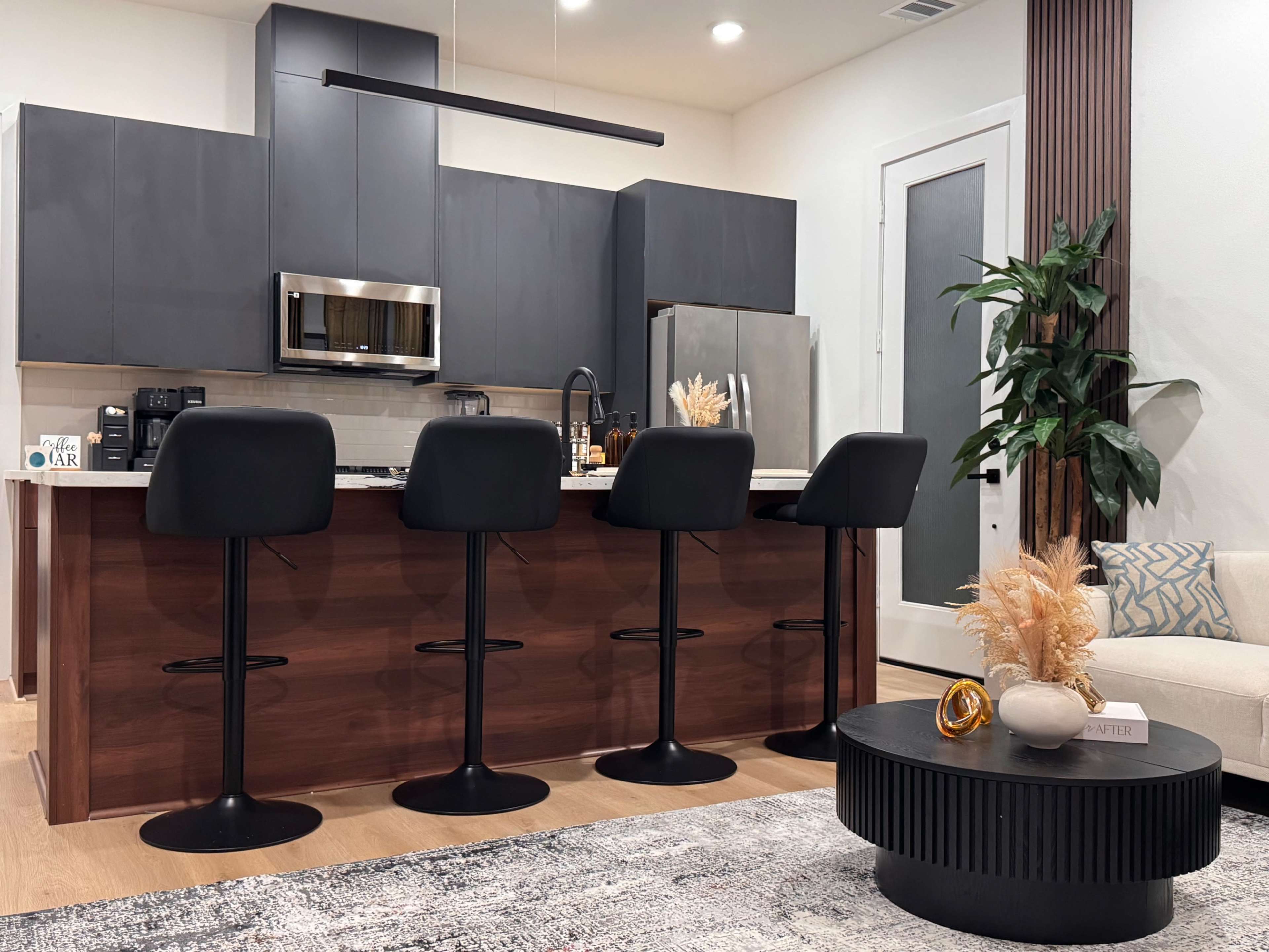 A modern kitchen features a dark cabinetry, stainless steel appliances, and four black bar stools at a wooden island, with a decorative plant and coffee table in the foreground.