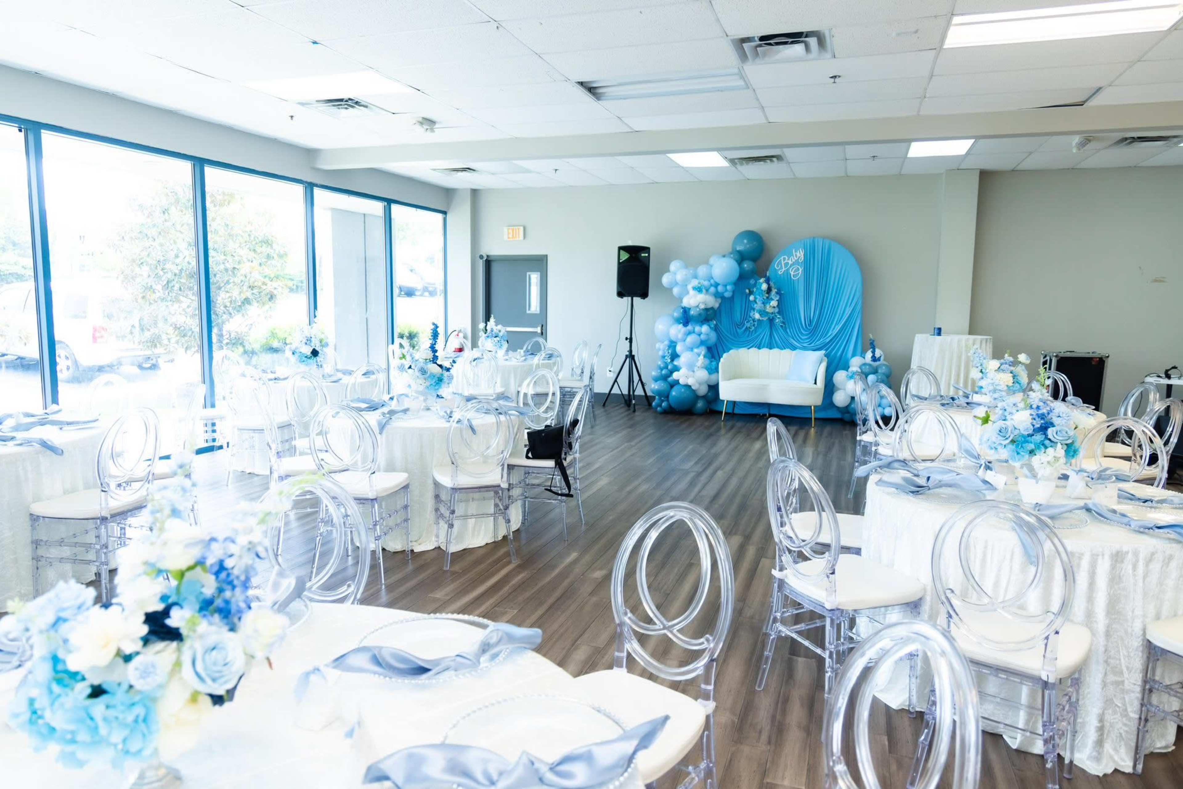 The room is set up for an event with circular tables adorned in white and blue, clear chairs, and a decorative backdrop featuring balloons and a sofa.