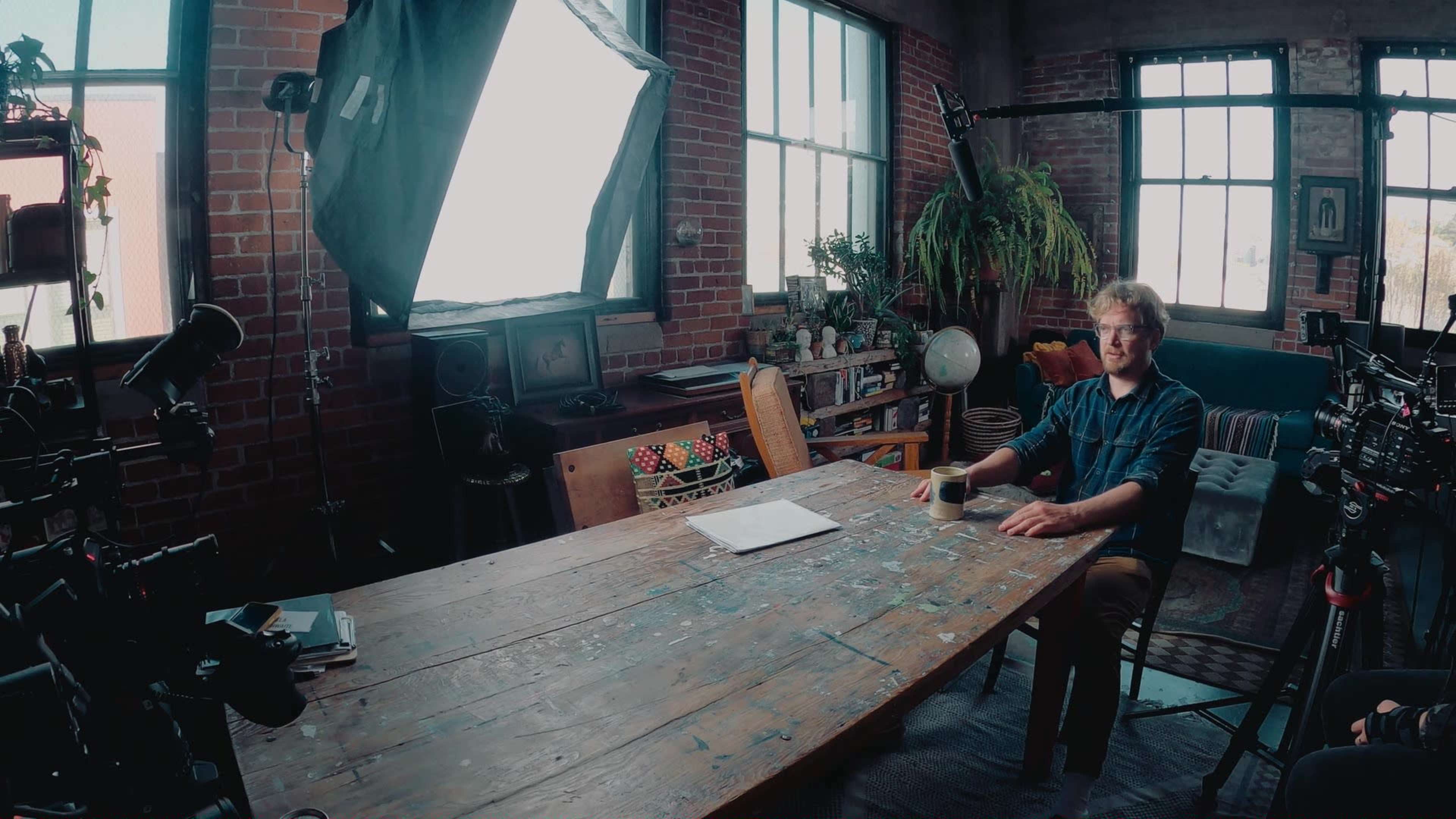 A man sits at a large, weathered wooden table in a studio filled with plants and equipment, with large windows letting in natural light.