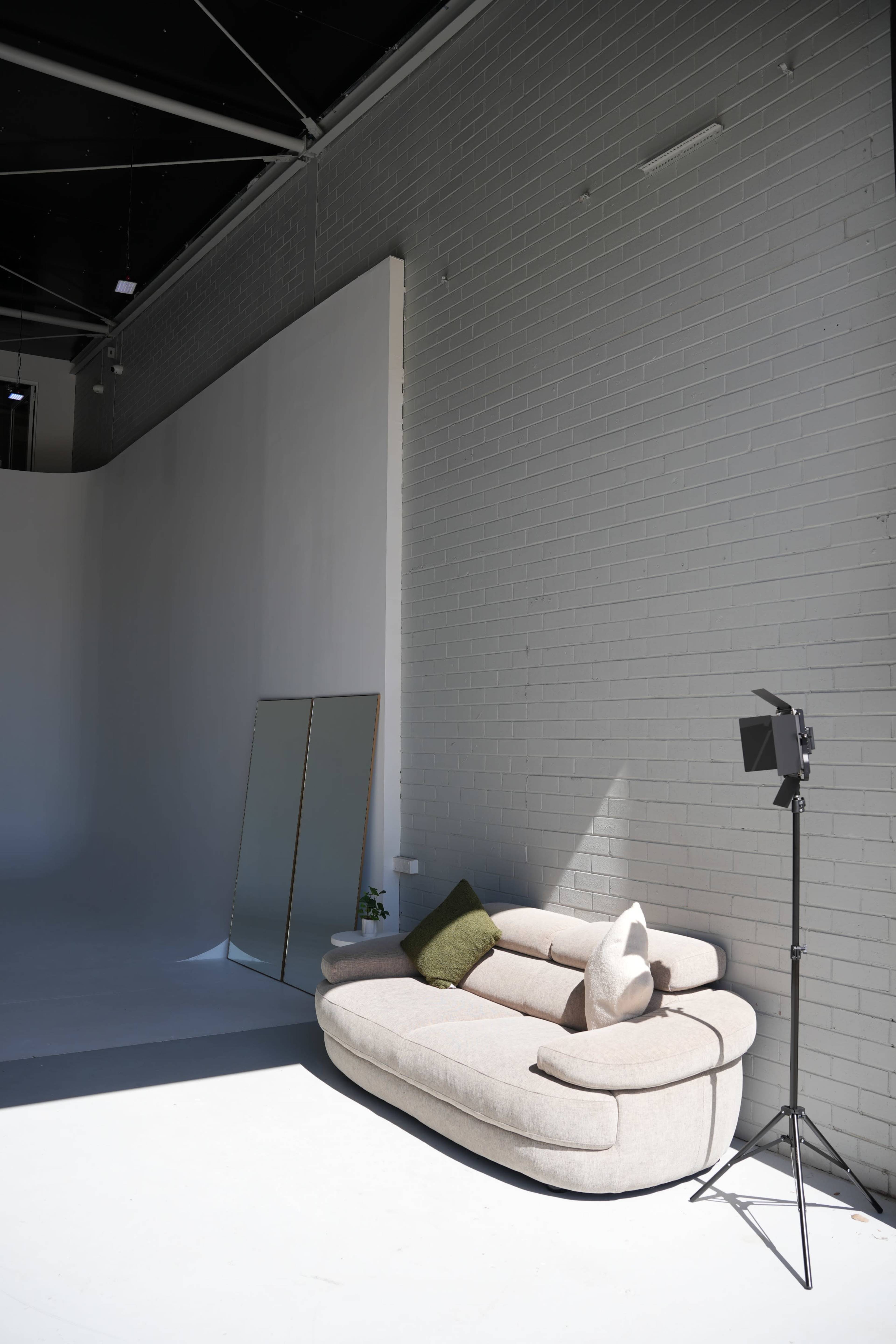 A light-filled studio features a curved beige sofa with two pillows, a tall mirror, and a floor lamp against a plain white wall.