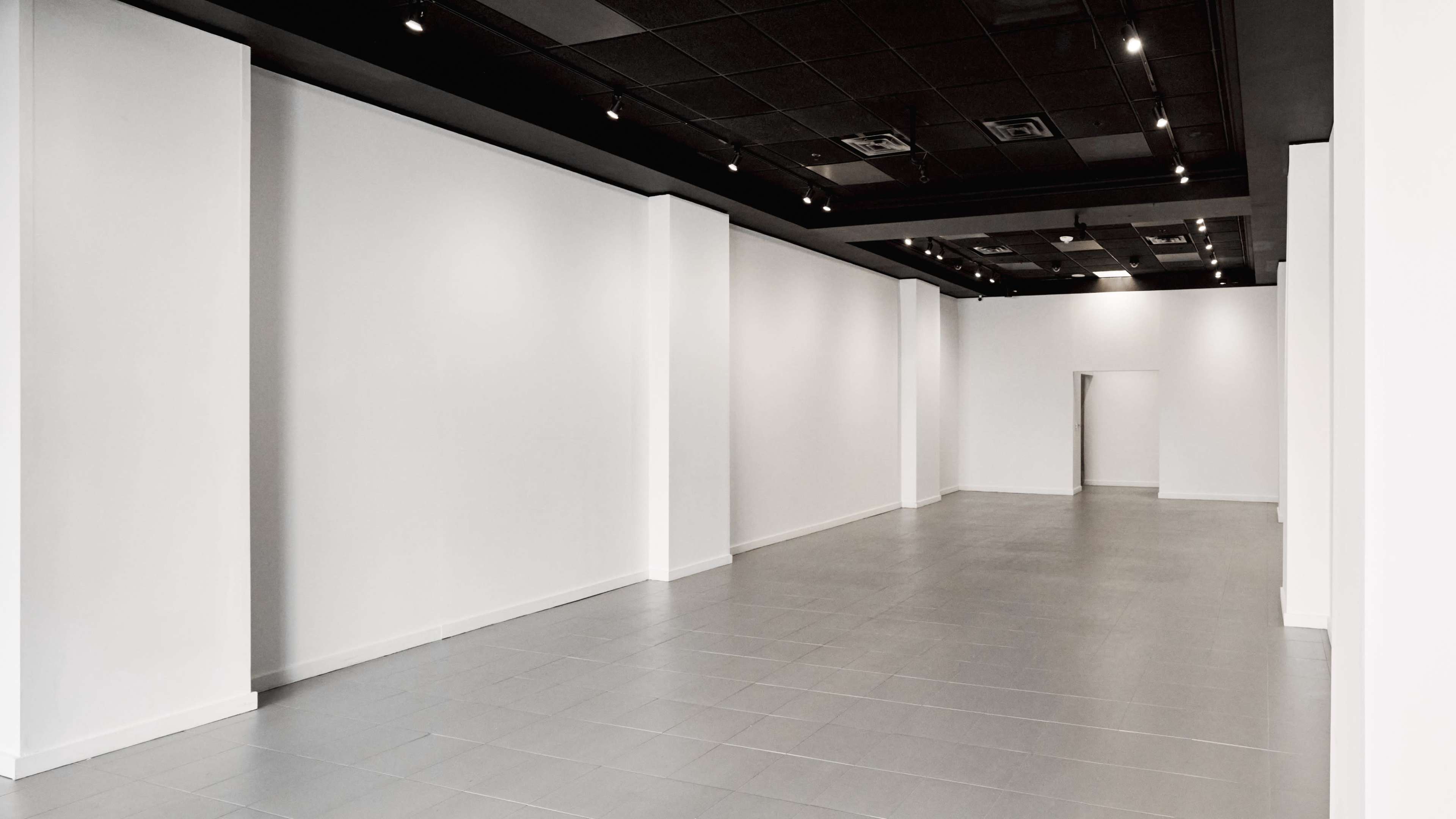 The image shows a spacious, empty interior room with white walls and gray tiled flooring, illuminated by overhead lights.