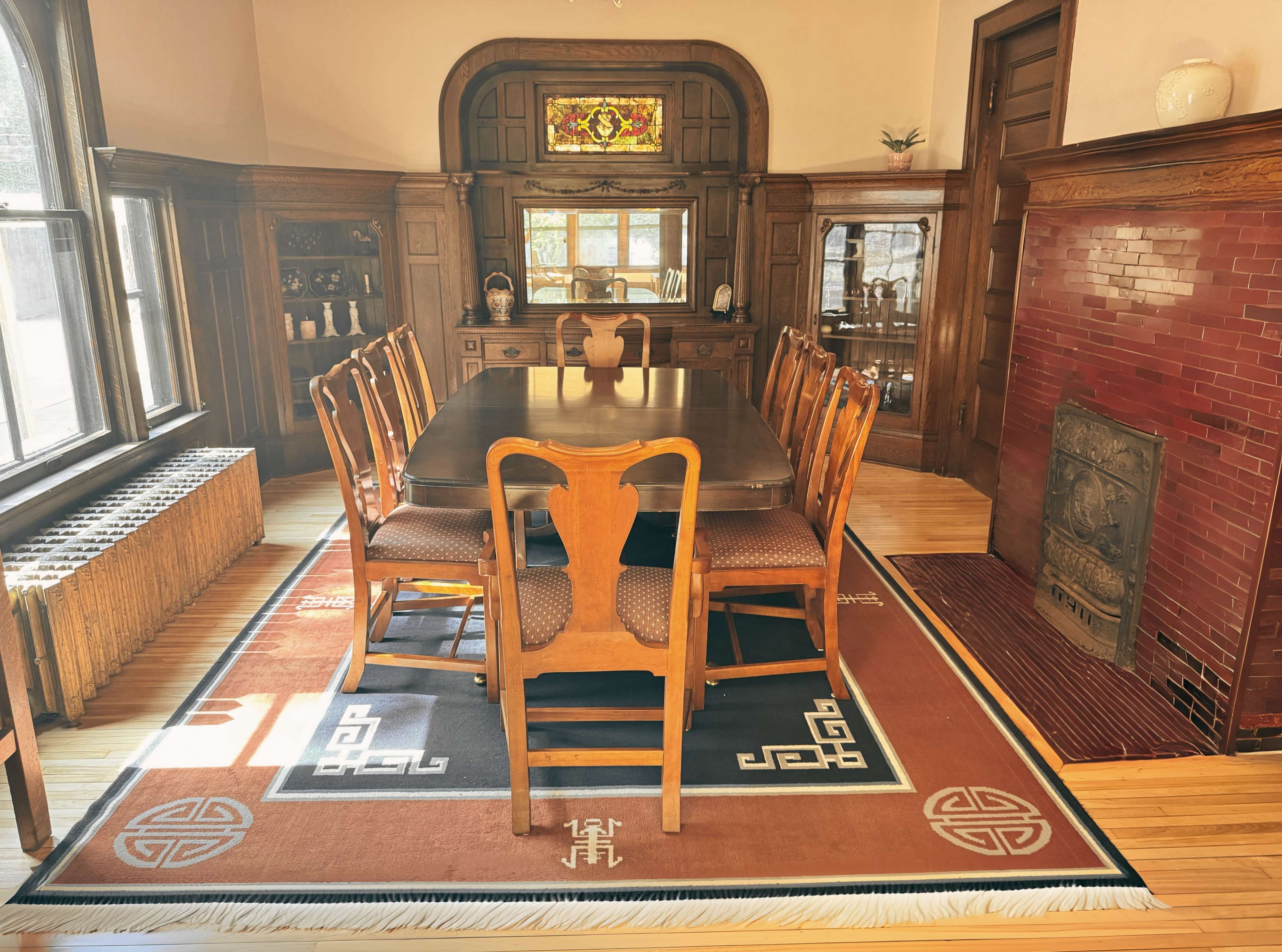 The image shows a spacious dining room featuring a large wooden table surrounded by eight chairs, an ornate sideboard, and a decorative fireplace with red tiles.