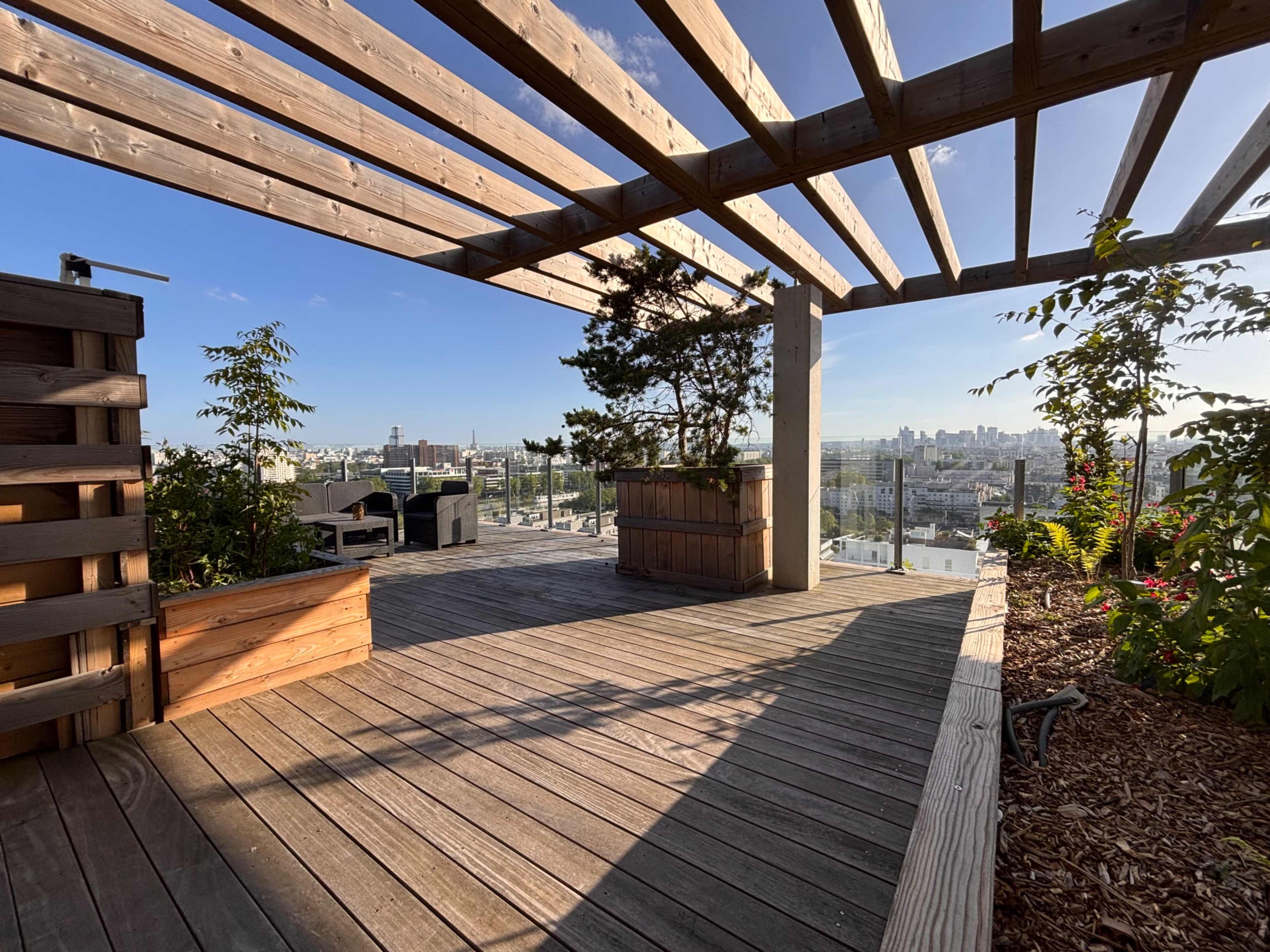An outdoor wooden terrace features a pergola, potted plants, and a view of a city skyline in the background.