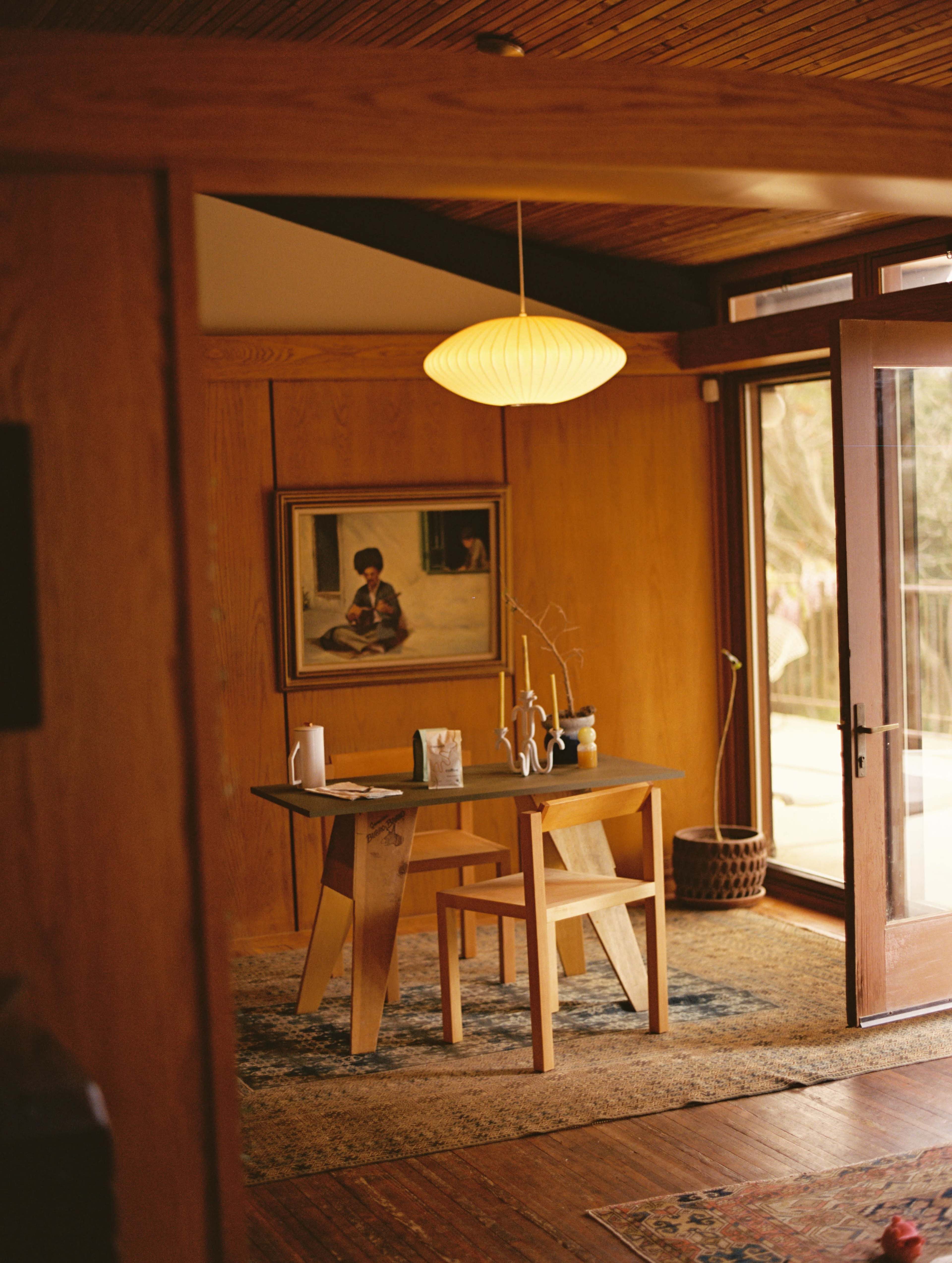 A cozy interior features a wooden dining table with two chairs, illuminated by a pendant light, surrounded by warm wood paneling and large glass doors.
