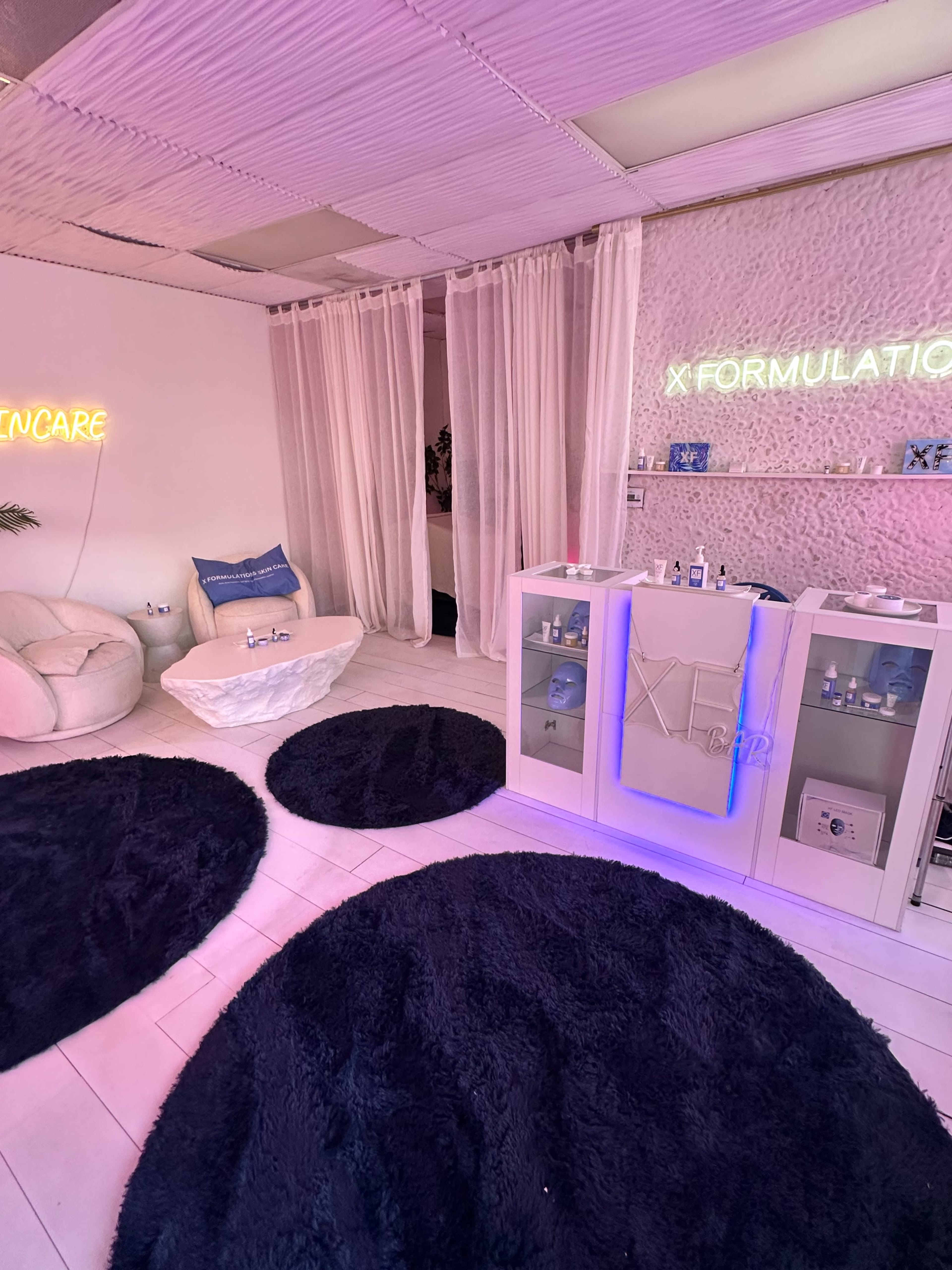 The image shows a minimalist spa room with soft lighting, featuring white walls, plush seating, decorative rugs, and a display counter with skincare products.