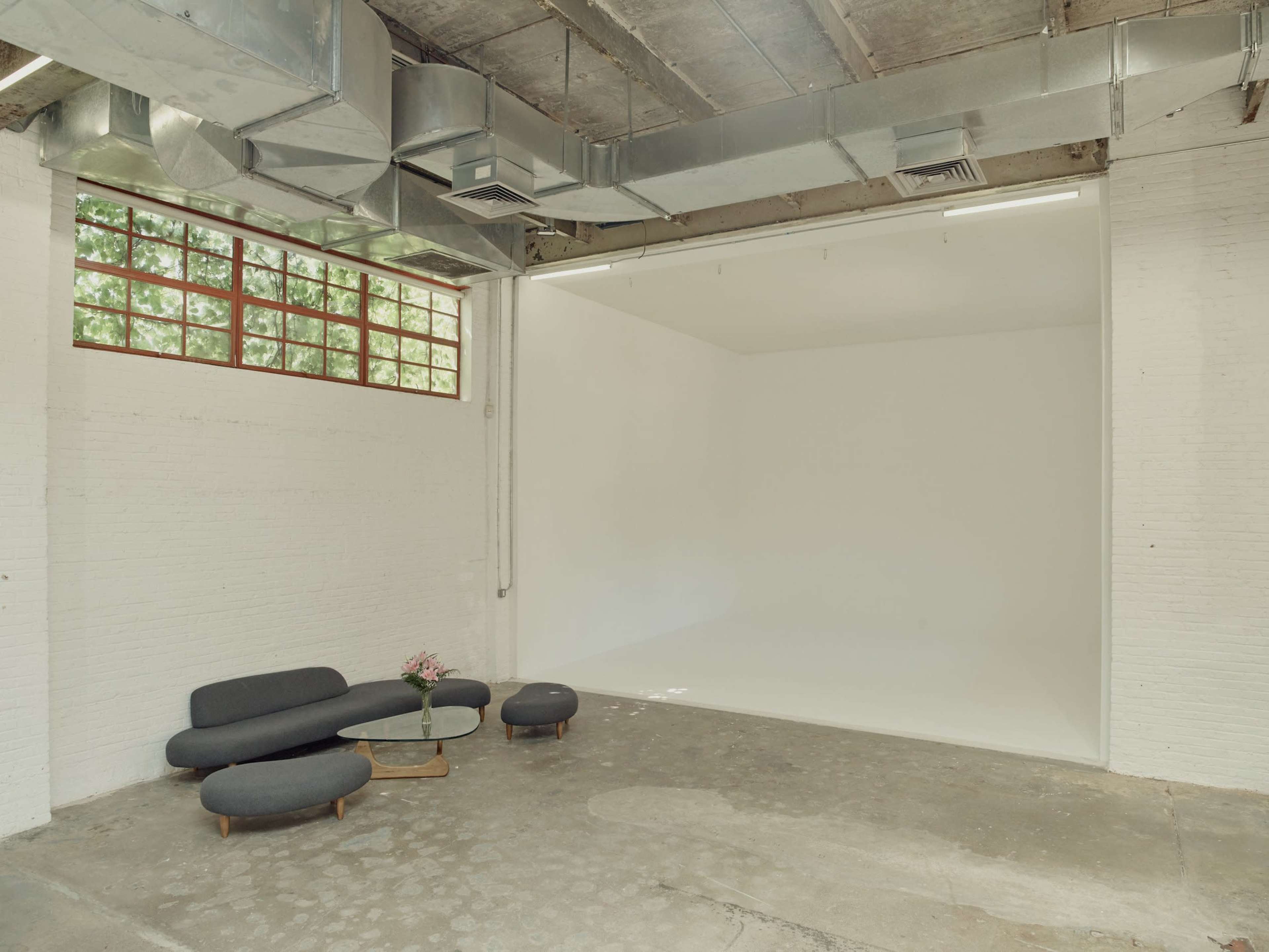 The image shows a spacious, minimalist studio with a gray couch and coffee table, featuring a large blank wall illuminated by natural light from a window.