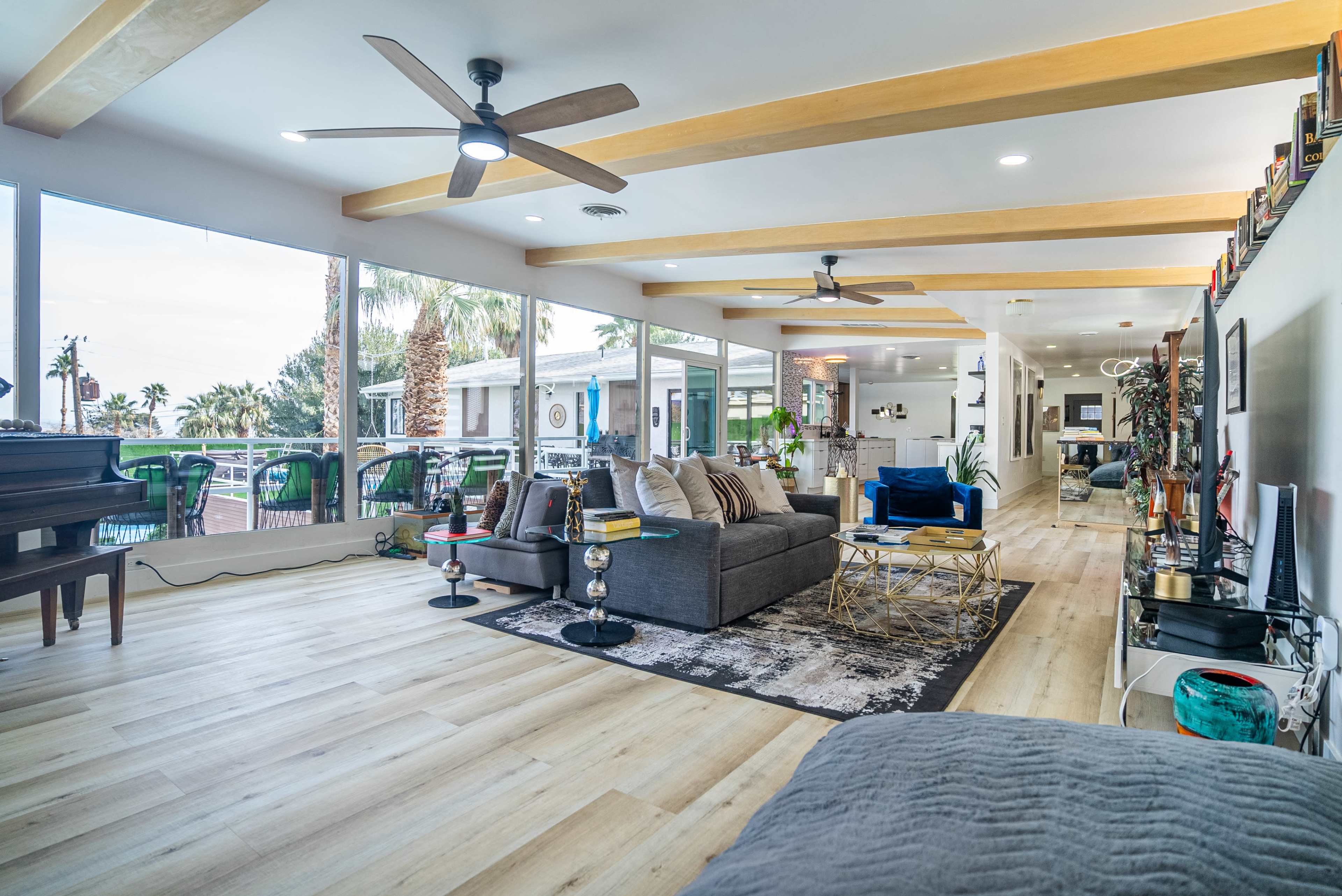 The image shows a modern open-concept living room with large windows, wooden beams on the ceiling, and a mix of contemporary furniture including a gray sofa and a piano.