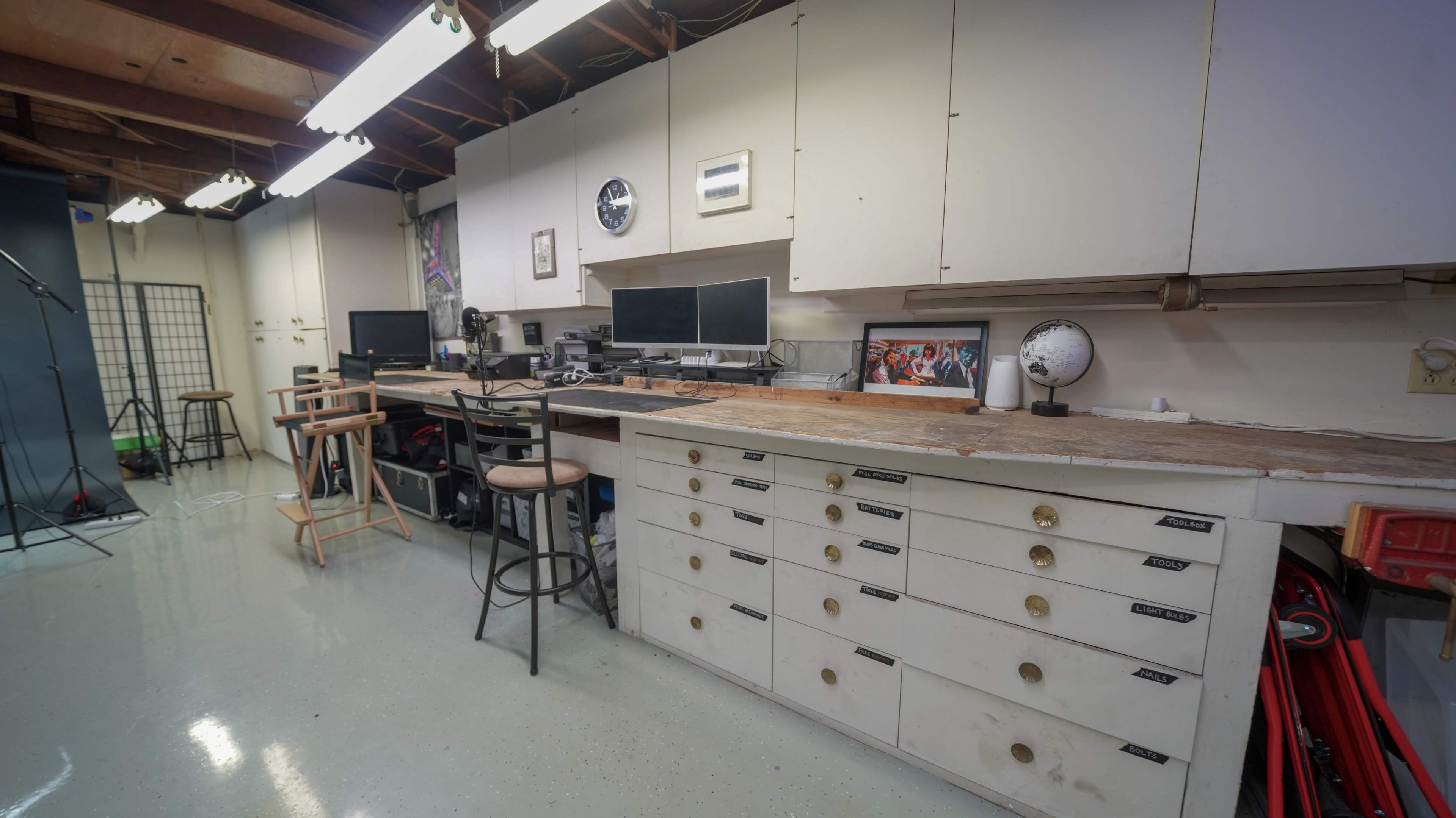 A well-organized workshop with multiple desks and computer monitors, featuring a long workbench with drawers and stools.