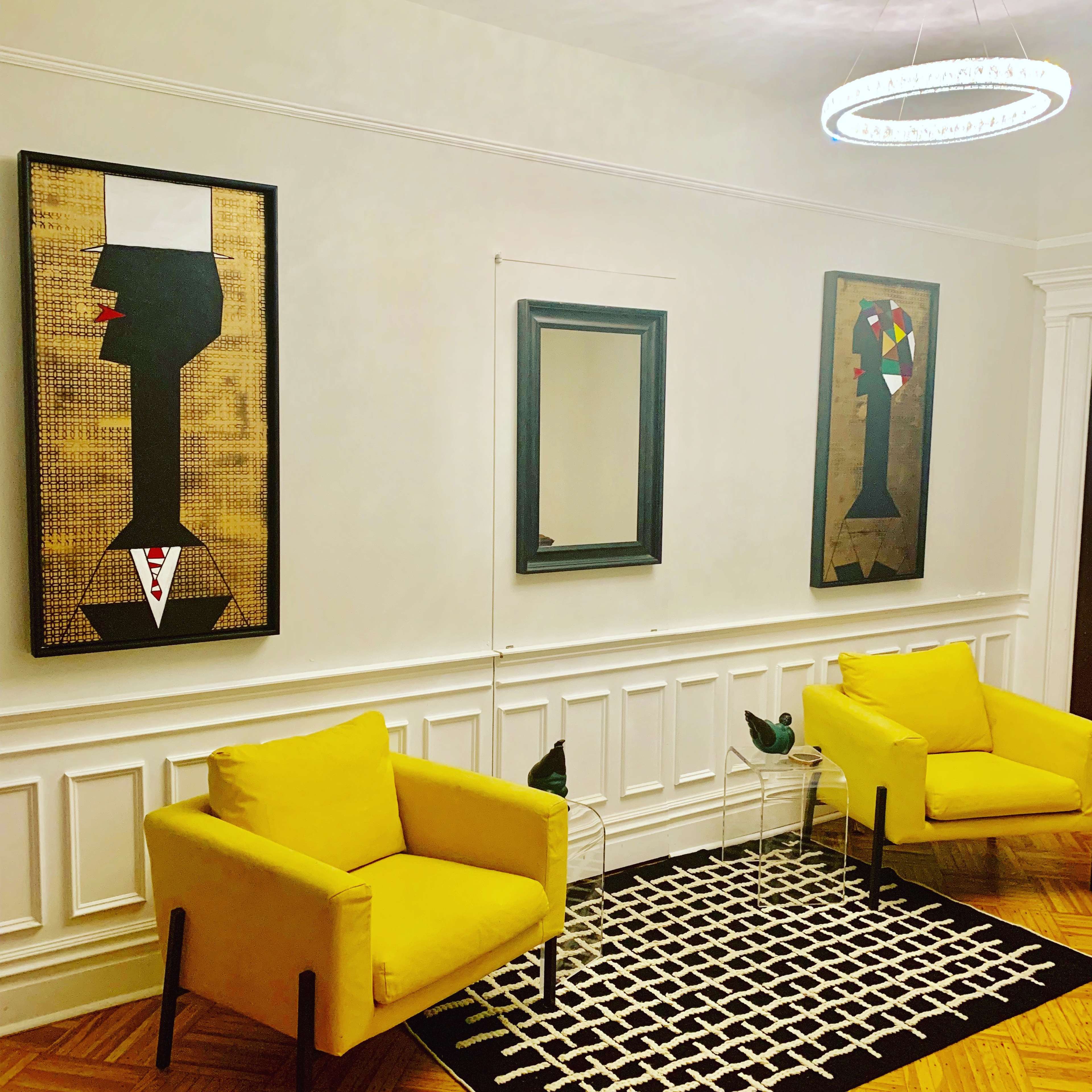 A room featuring two yellow chairs, a patterned rug, and three framed artworks on the walls, including a mirror.