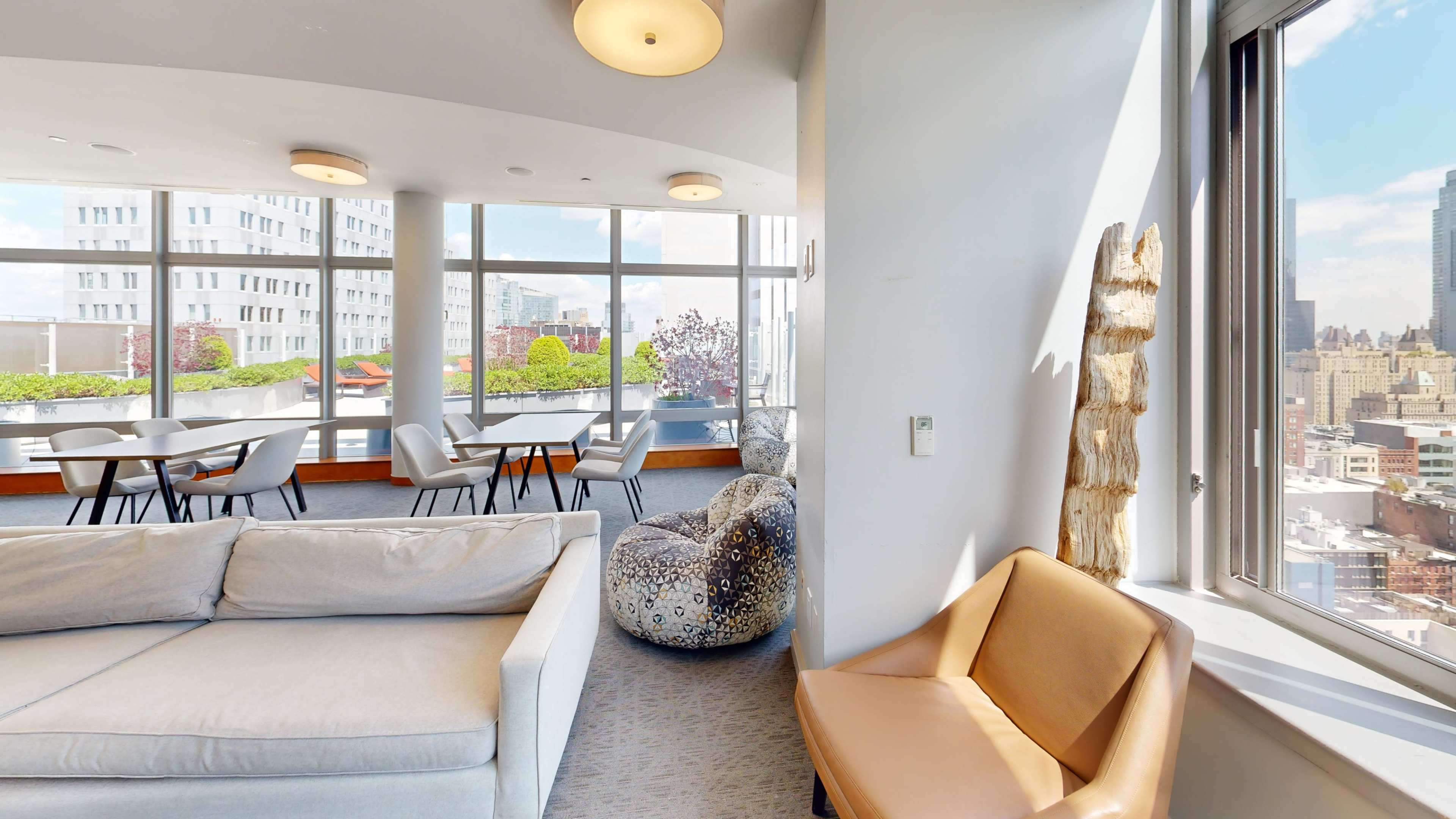 A modern interior space featuring a light-colored sofa, a brown chair, bean bag seating, and a panoramic view of the city through large windows.