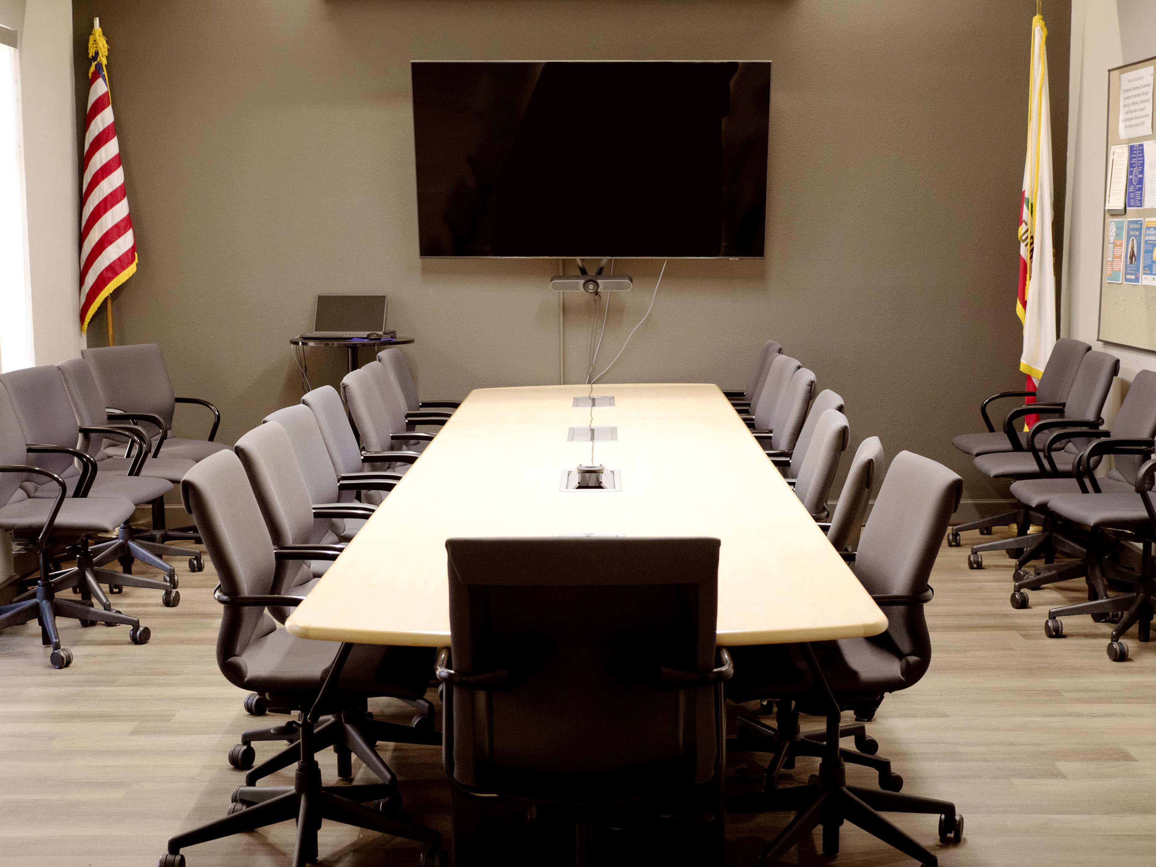The image shows a large conference room with a long rectangular table surrounded by gray chairs, featuring a large wall-mounted screen at one end.