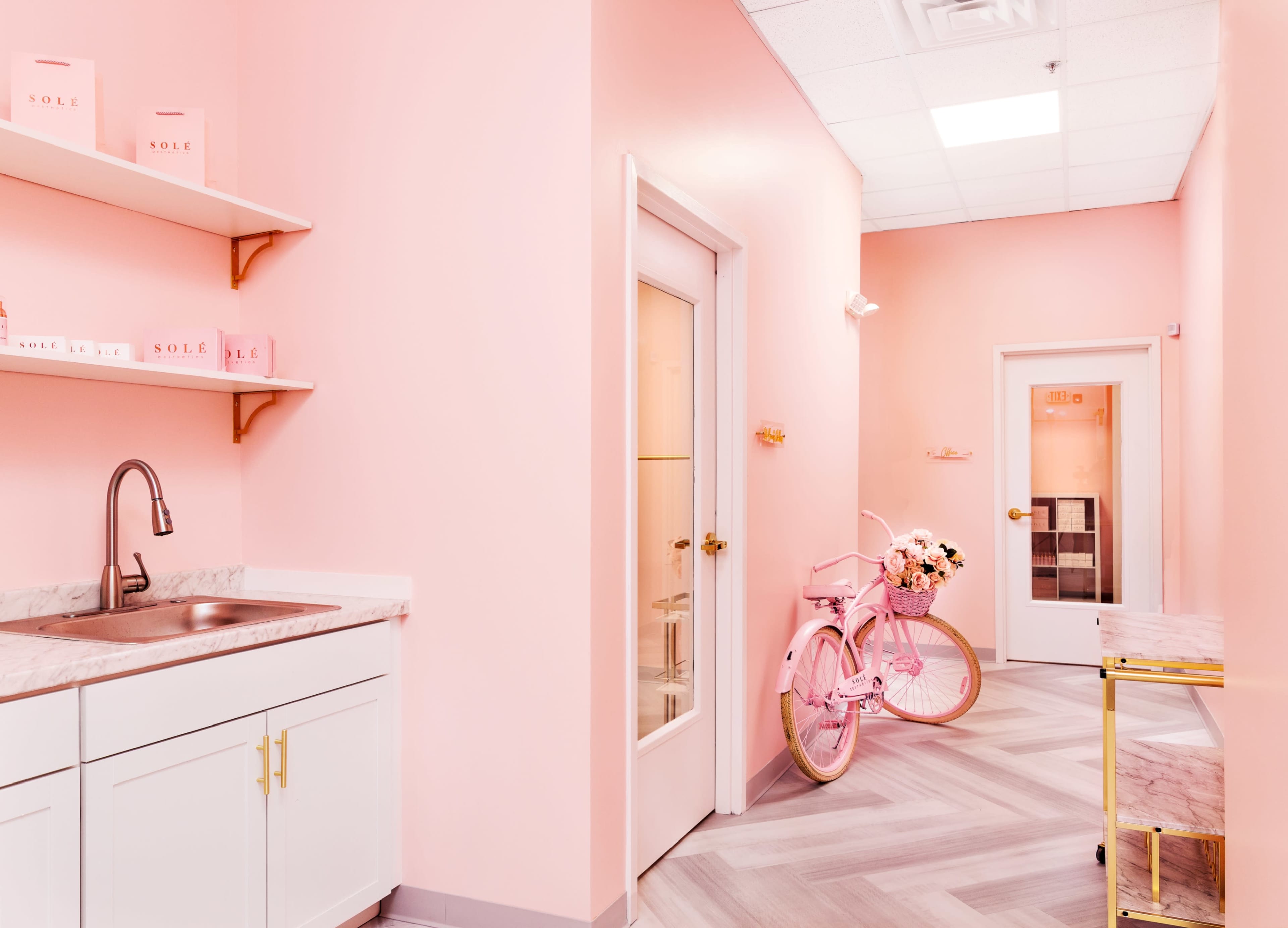 The image shows a bright pink hallway featuring a vintage pink bicycle with flowers, a sink area, and shelves with decorative boxes.