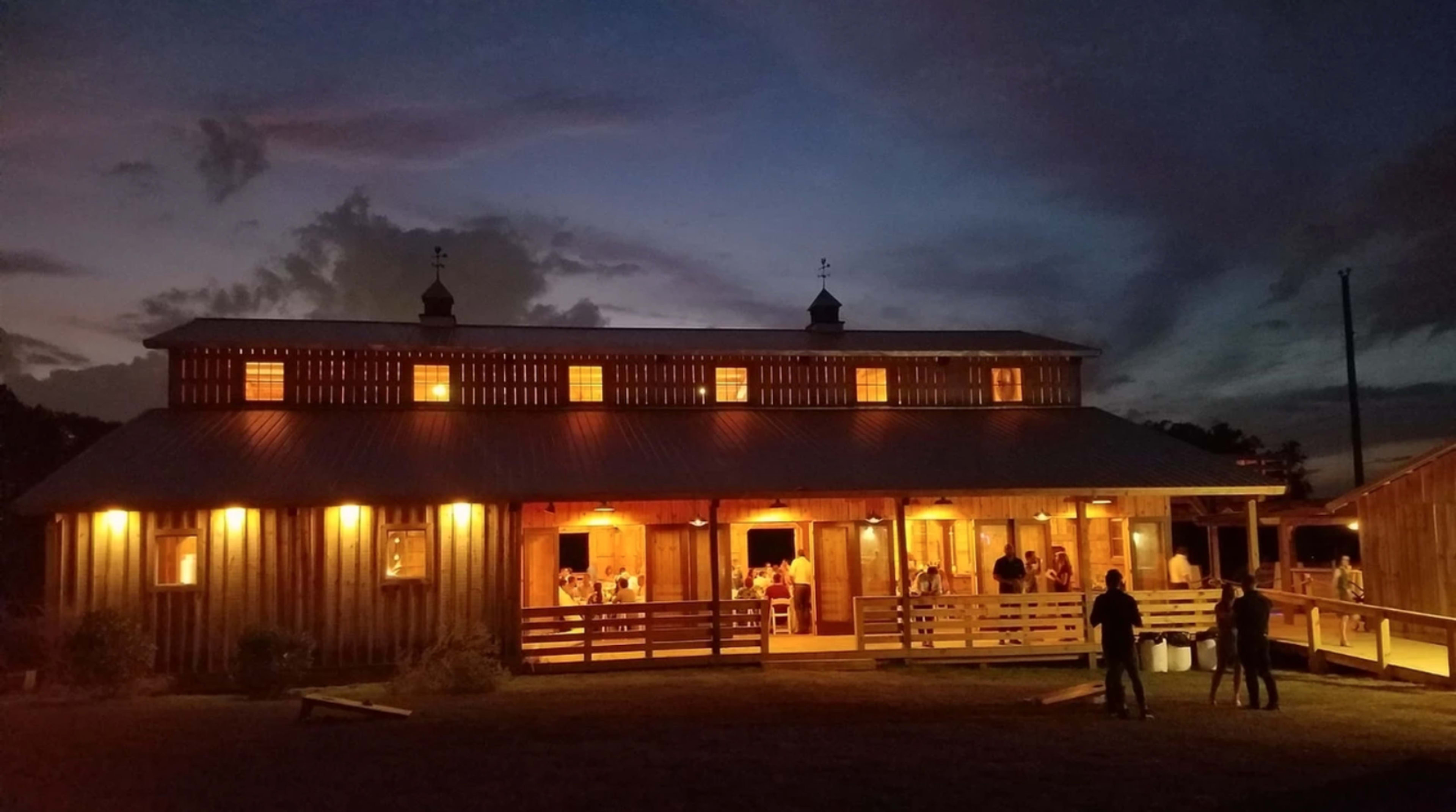 A wooden barn illuminated by warm lights stands against a twilight sky with people gathered on its porch.