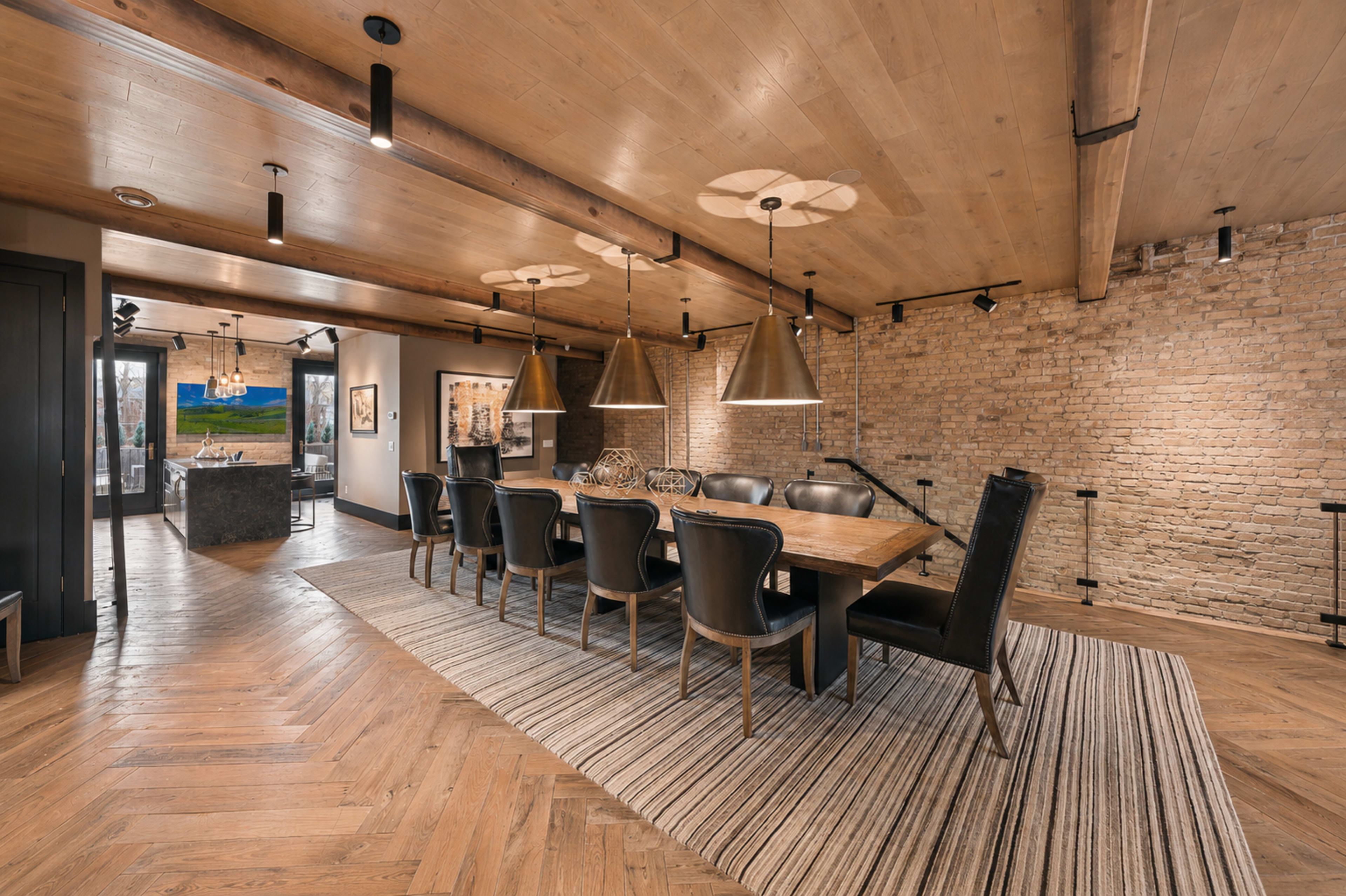 The image shows a well-lit dining area with a long wooden table surrounded by black leather chairs, featuring exposed brick walls and pendant lighting.