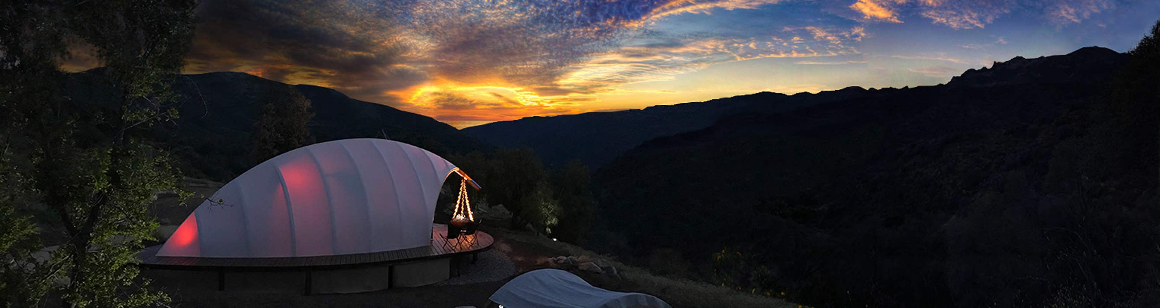 A dome-shaped structure sits on a hillside as the sun sets behind the mountains, casting colorful light across the sky.