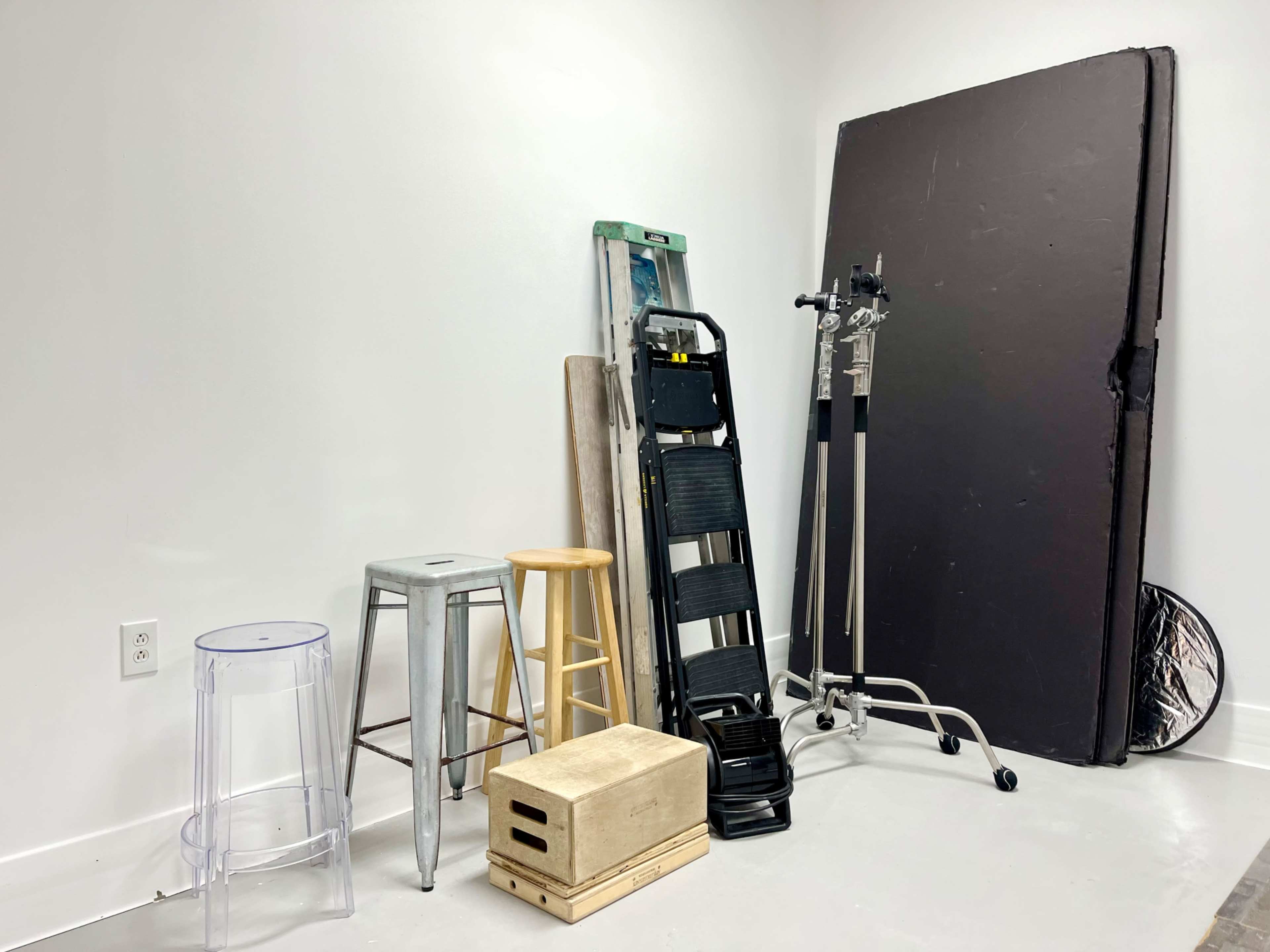 The image shows a collection of various items including stools, a ladder, and a large black board against a plain white wall.