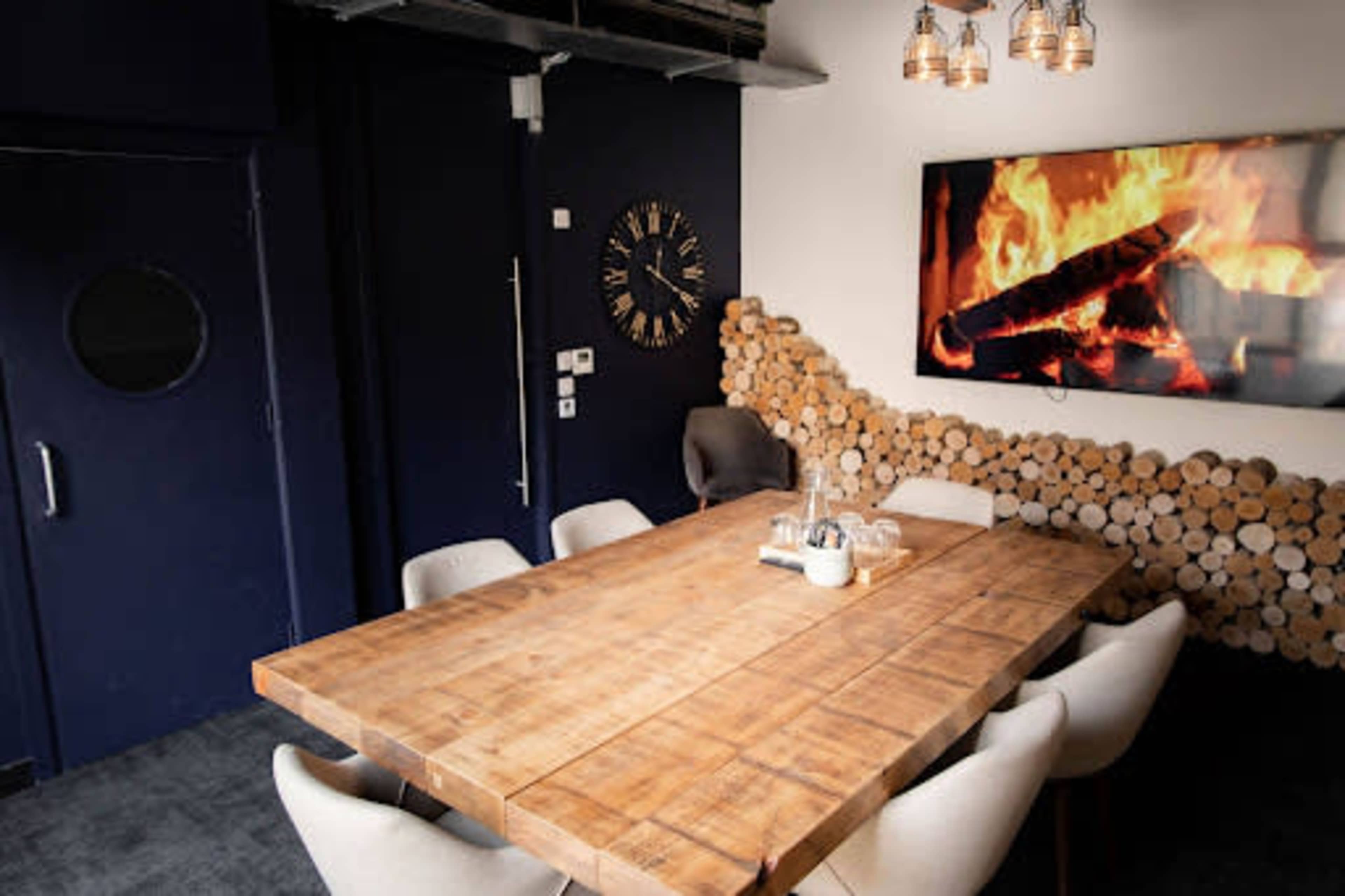 A wooden conference table surrounded by chairs is situated in a room with a dark wall and a television displaying a fire, alongside a feature wall made of stacked logs.