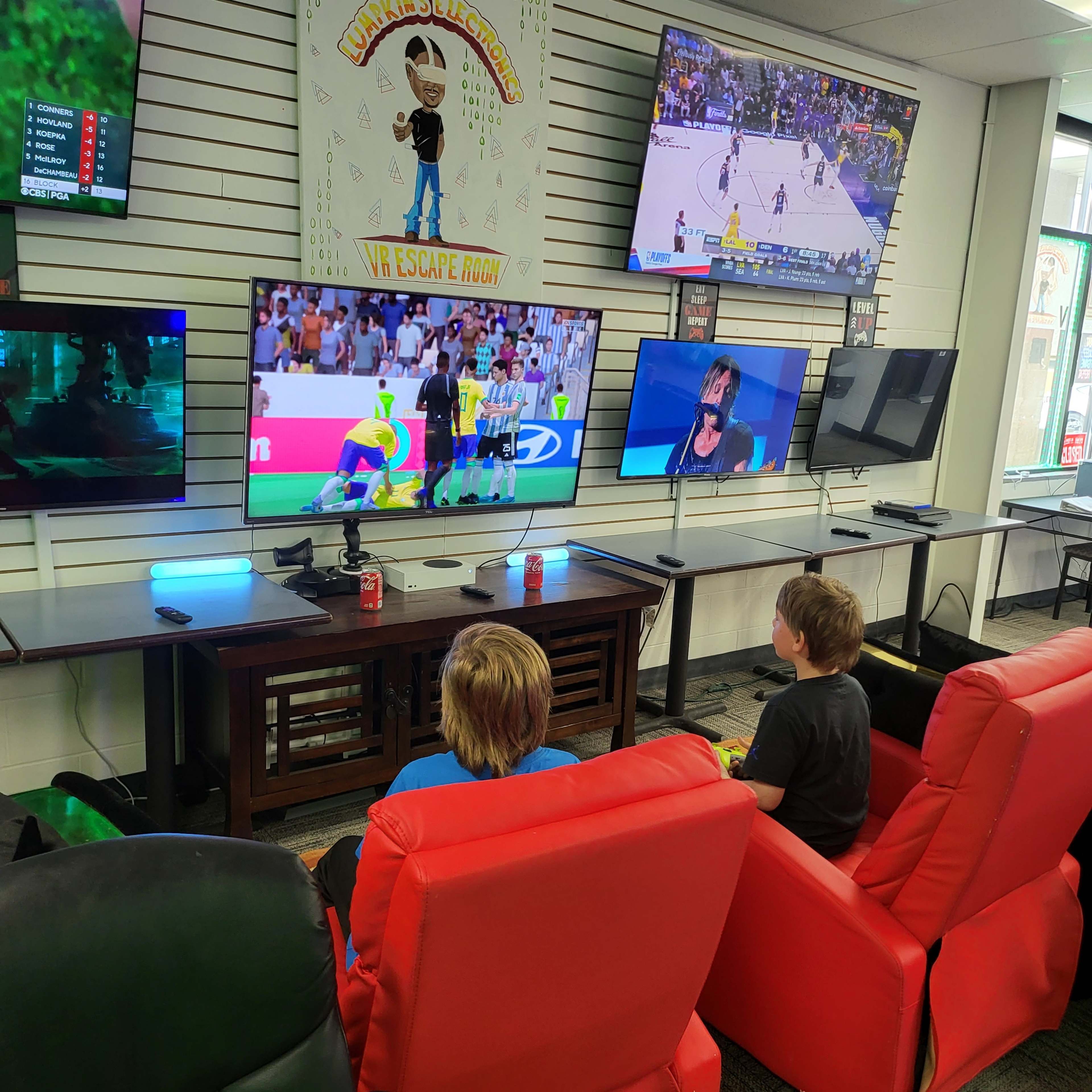 Two children sit in red chairs, watching multiple televisions displaying various sports and gaming content in a recreational area.