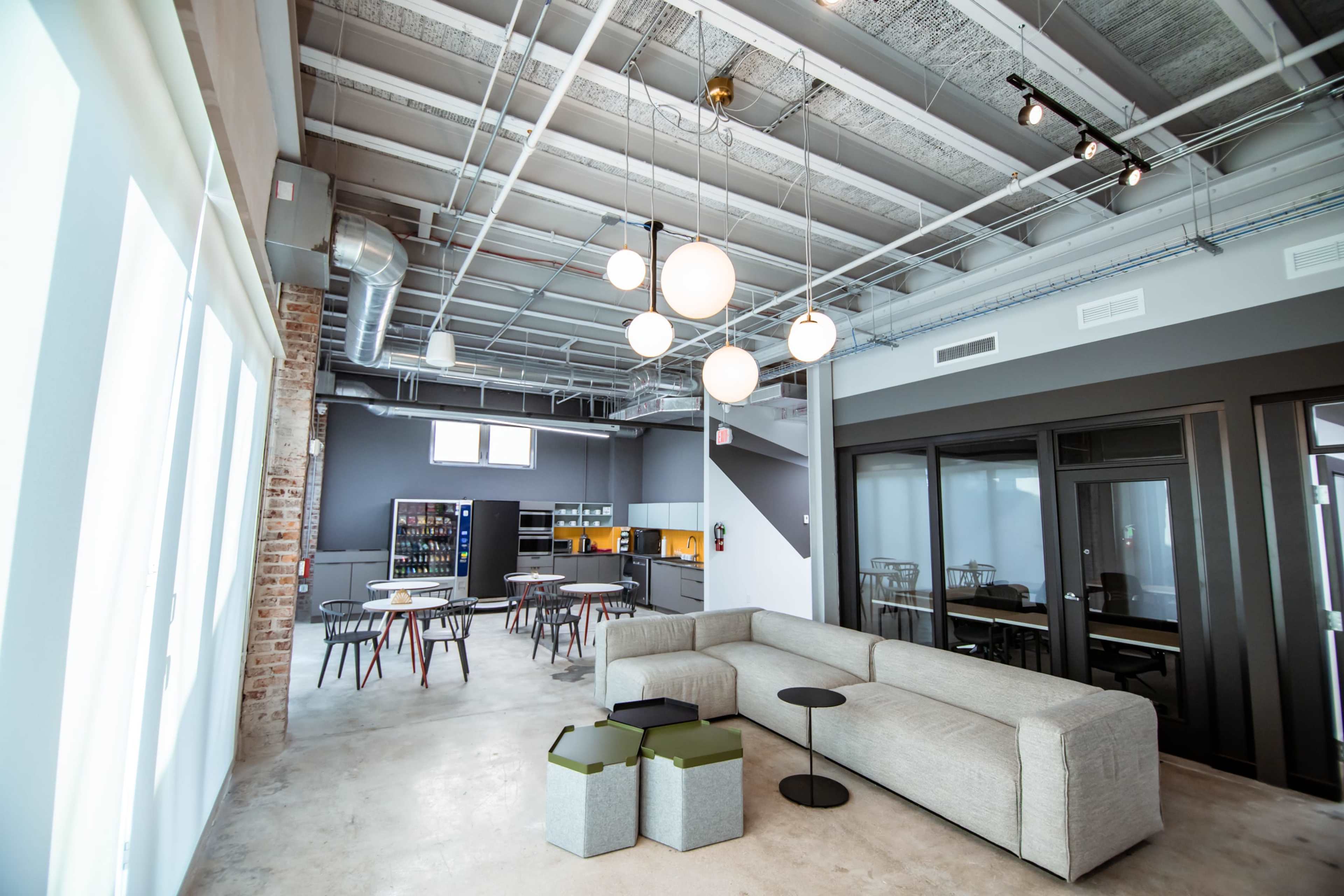 A modern open office space featuring a light-filled area with a grey sectional sofa, round tables, and a kitchenette in the background.