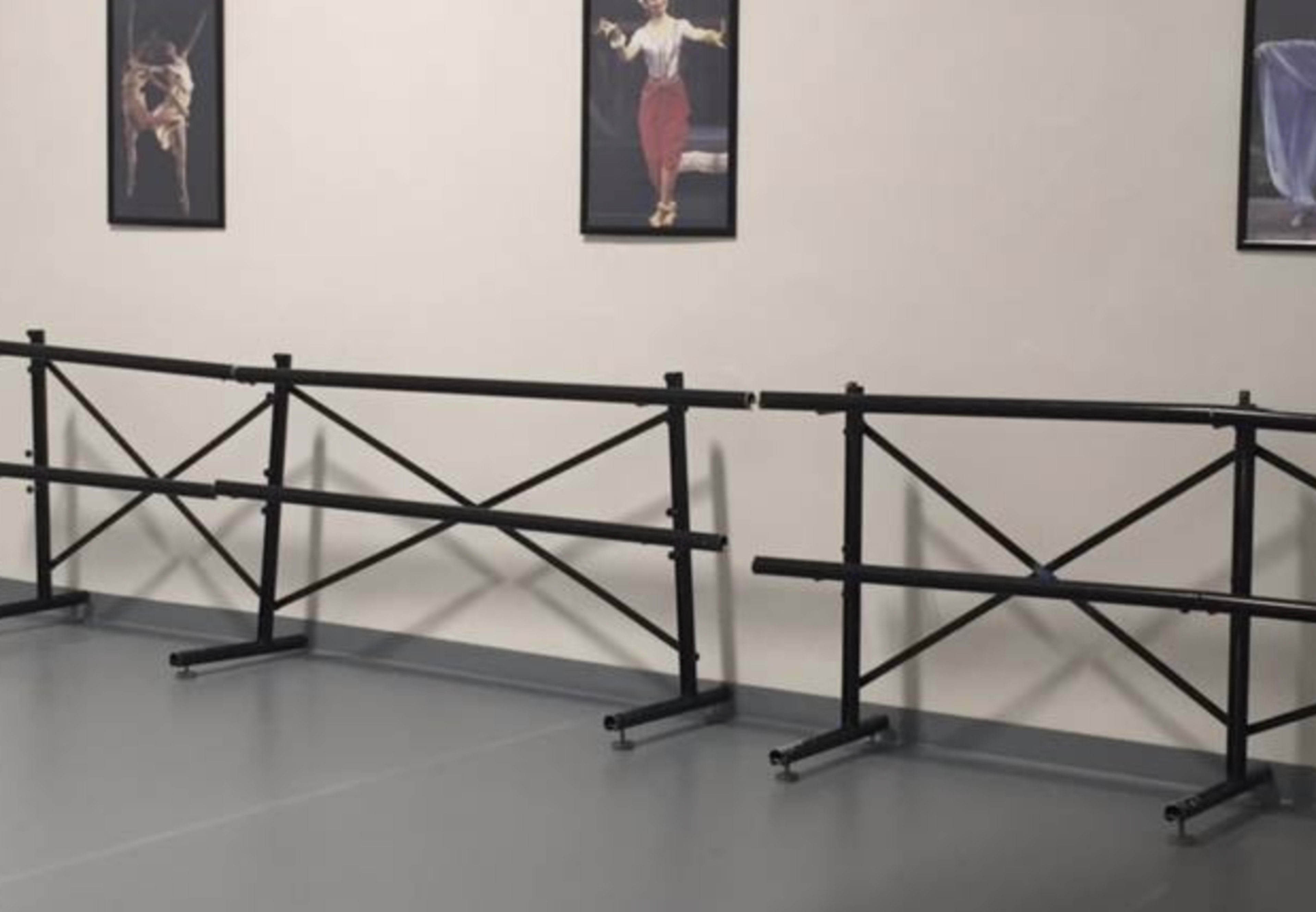 The image shows several black ballet barres positioned in a dance studio with framed dance-related artwork on the wall.
