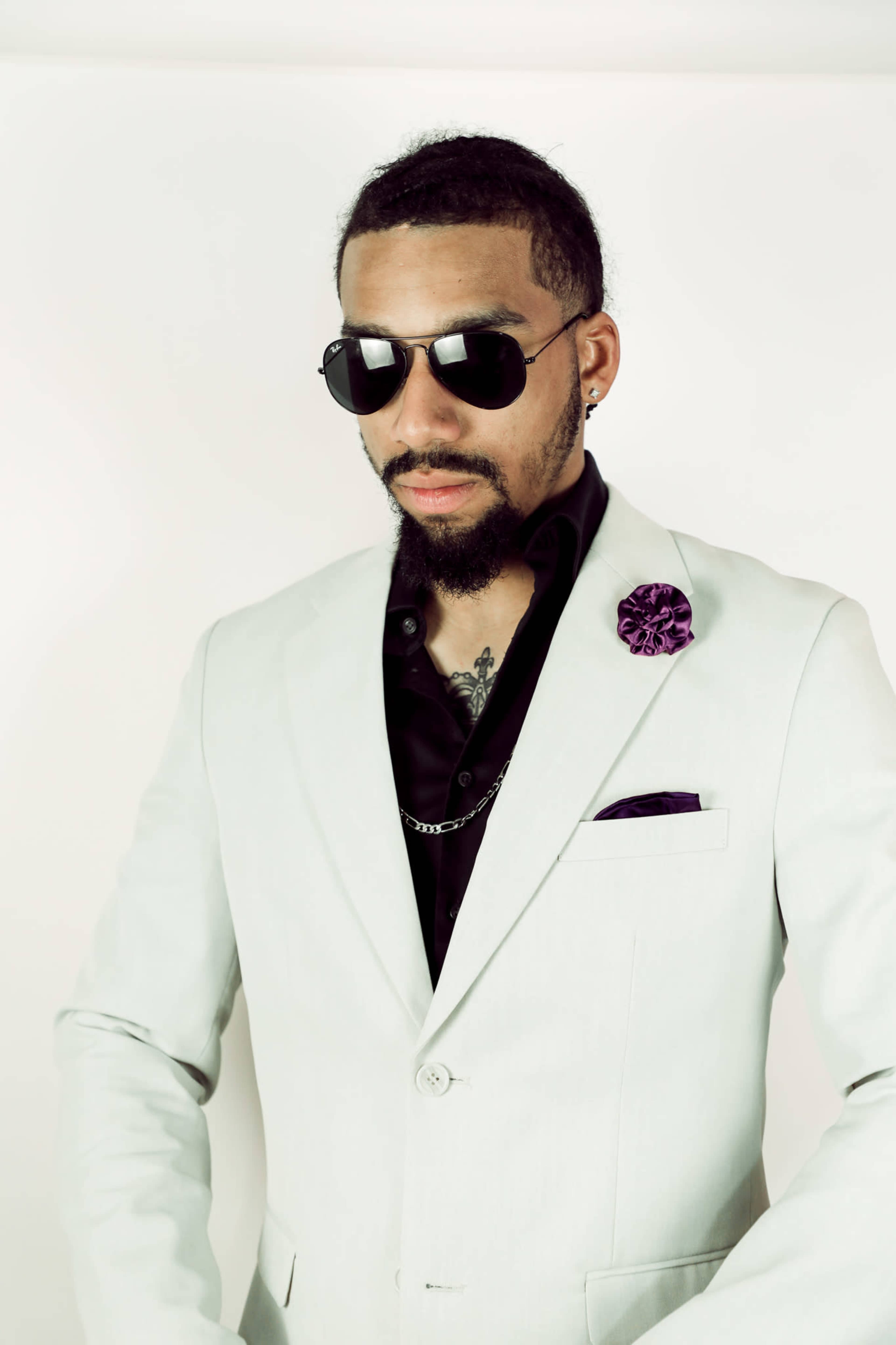 A man in a light-colored suit and black shirt poses confidently while wearing sunglasses and a purple lapel flower.