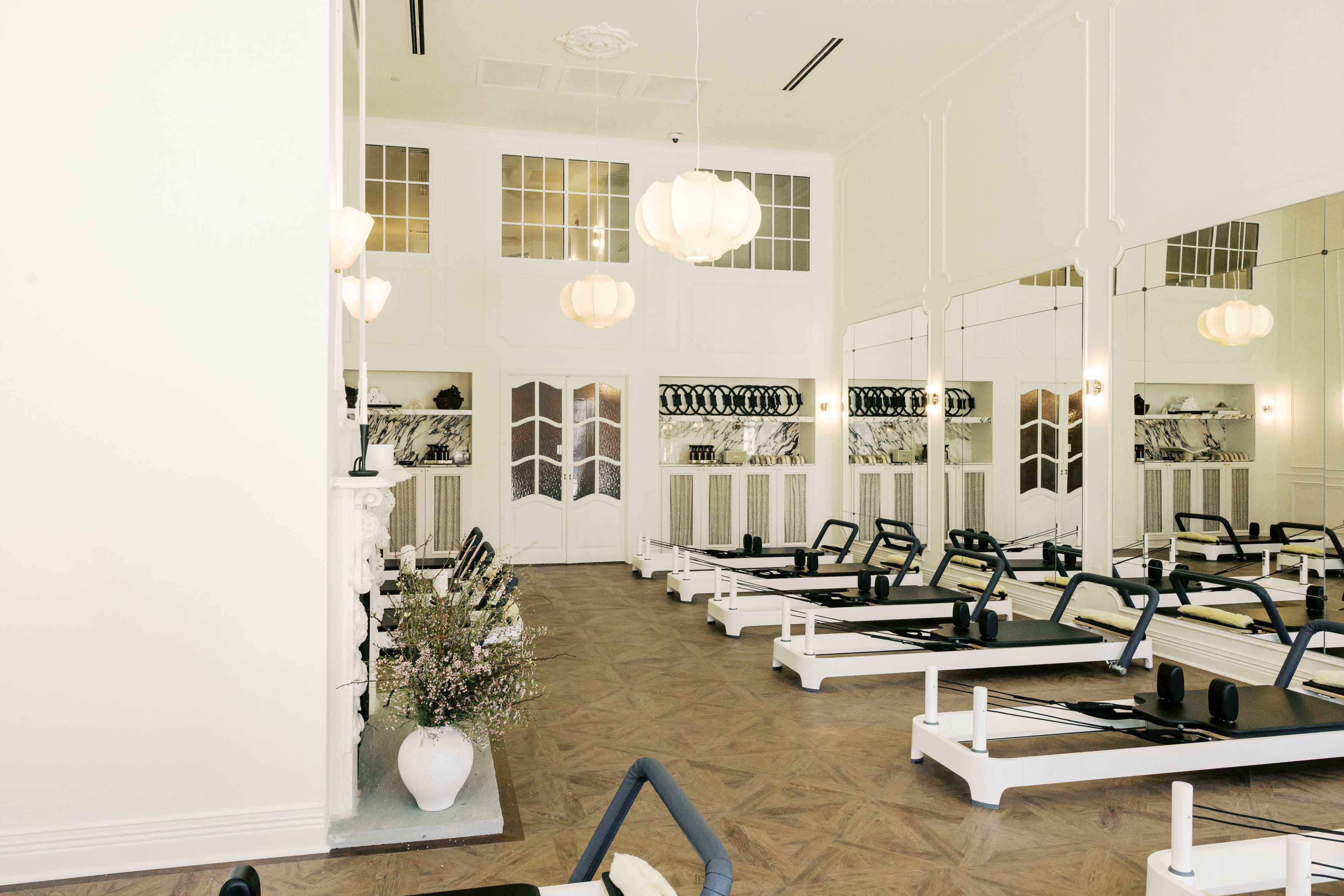The image shows a bright, modern Pilates studio featuring reformer machines, large mirrors, and elegant lighting fixtures.