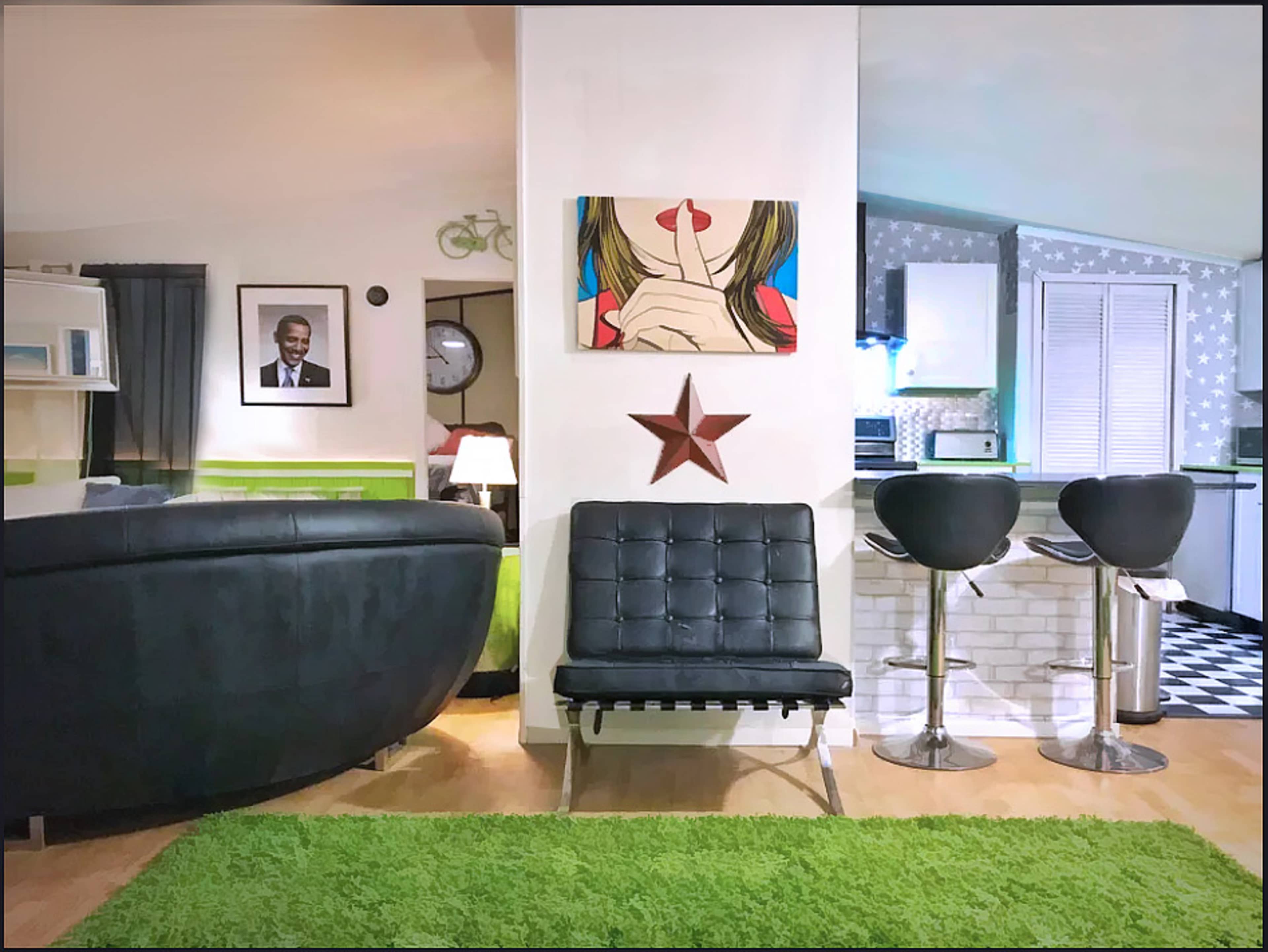 A modern living space featuring a black leather sofa, a green area rug, a star decoration on the wall, and a small bar area with two black stools.