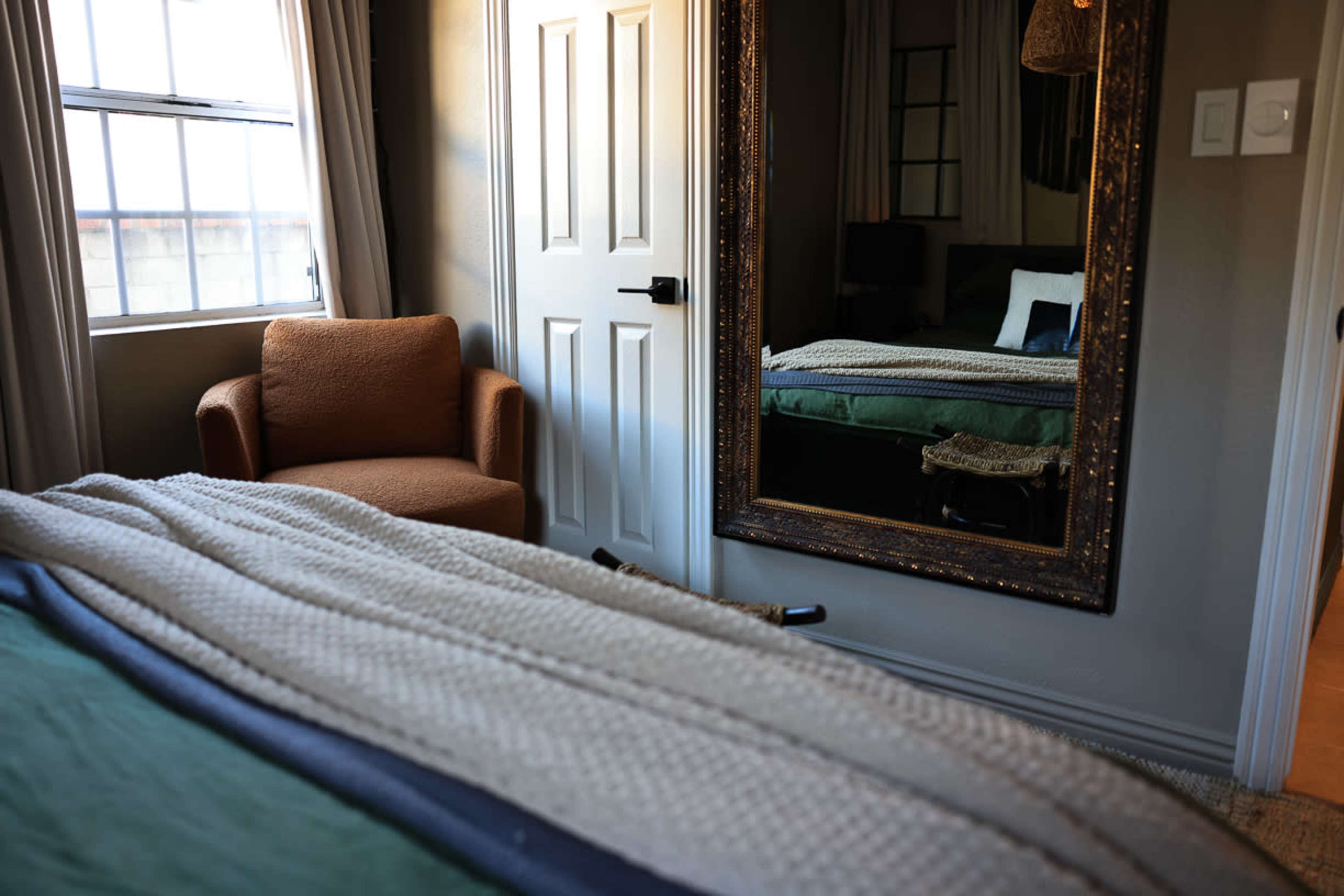 The image shows a cozy bedroom with a bed covered in a textured blanket, a brown armchair, and a large decorative mirror reflecting part of the room.