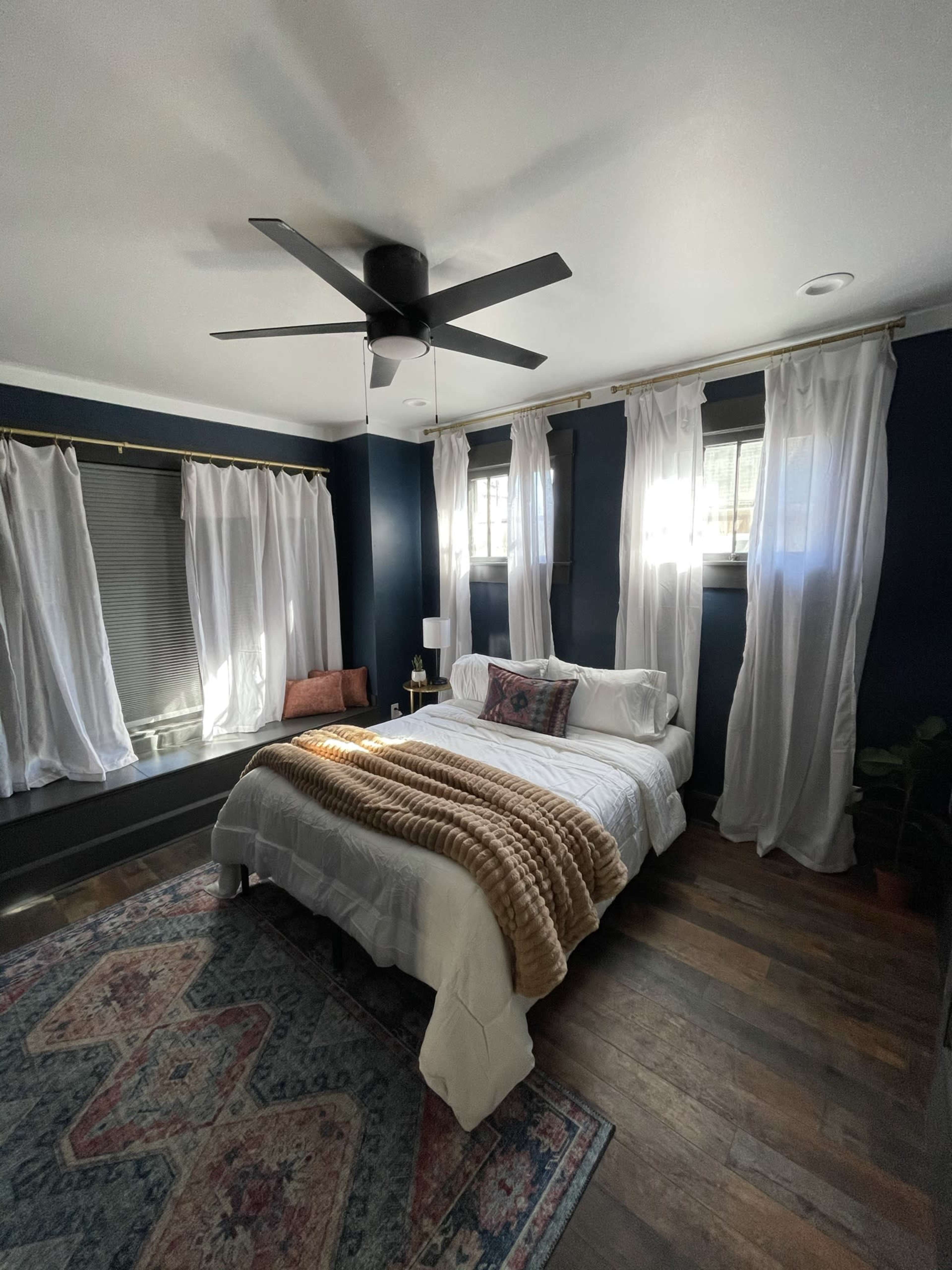 A well-lit bedroom featuring a bed with a white comforter, a decorative throw blanket, large windows with sheer curtains, and a ceiling fan.