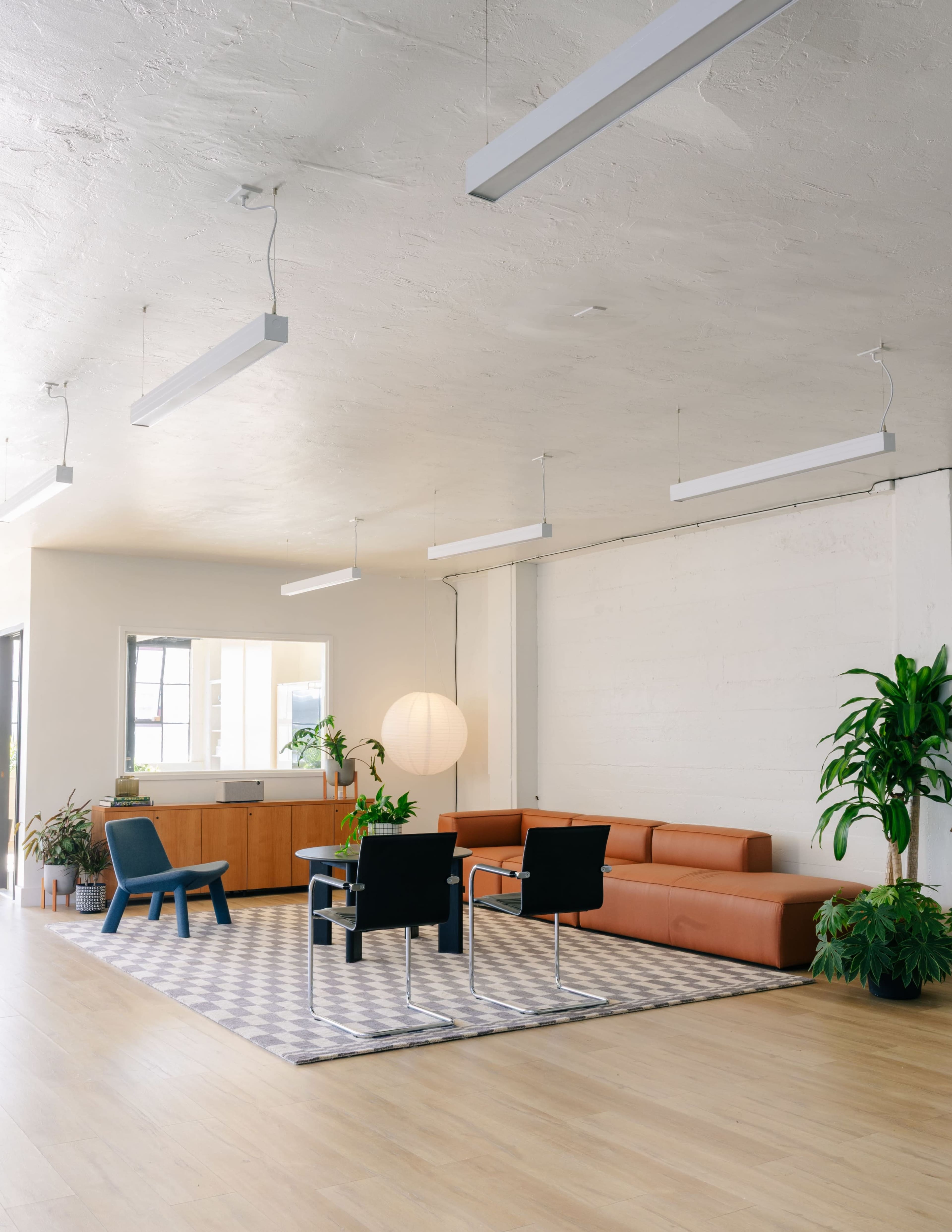 The image shows a modern, minimalist office space with a brown sectional sofa, two black chairs, a patterned rug, and large windows allowing natural light to enter.
