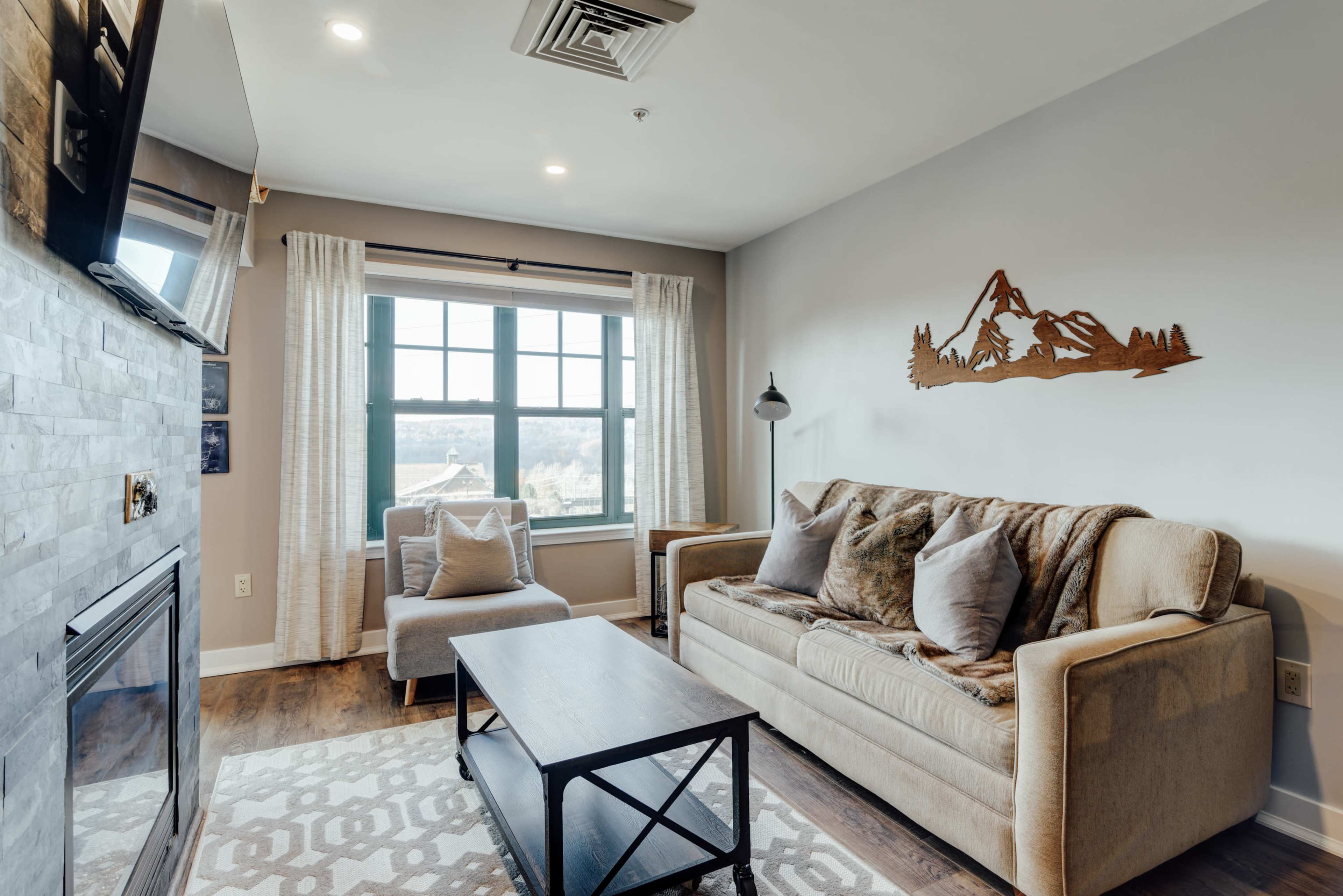 A living room features a sofa, a coffee table, and large windows with mountain-view artwork on the wall.