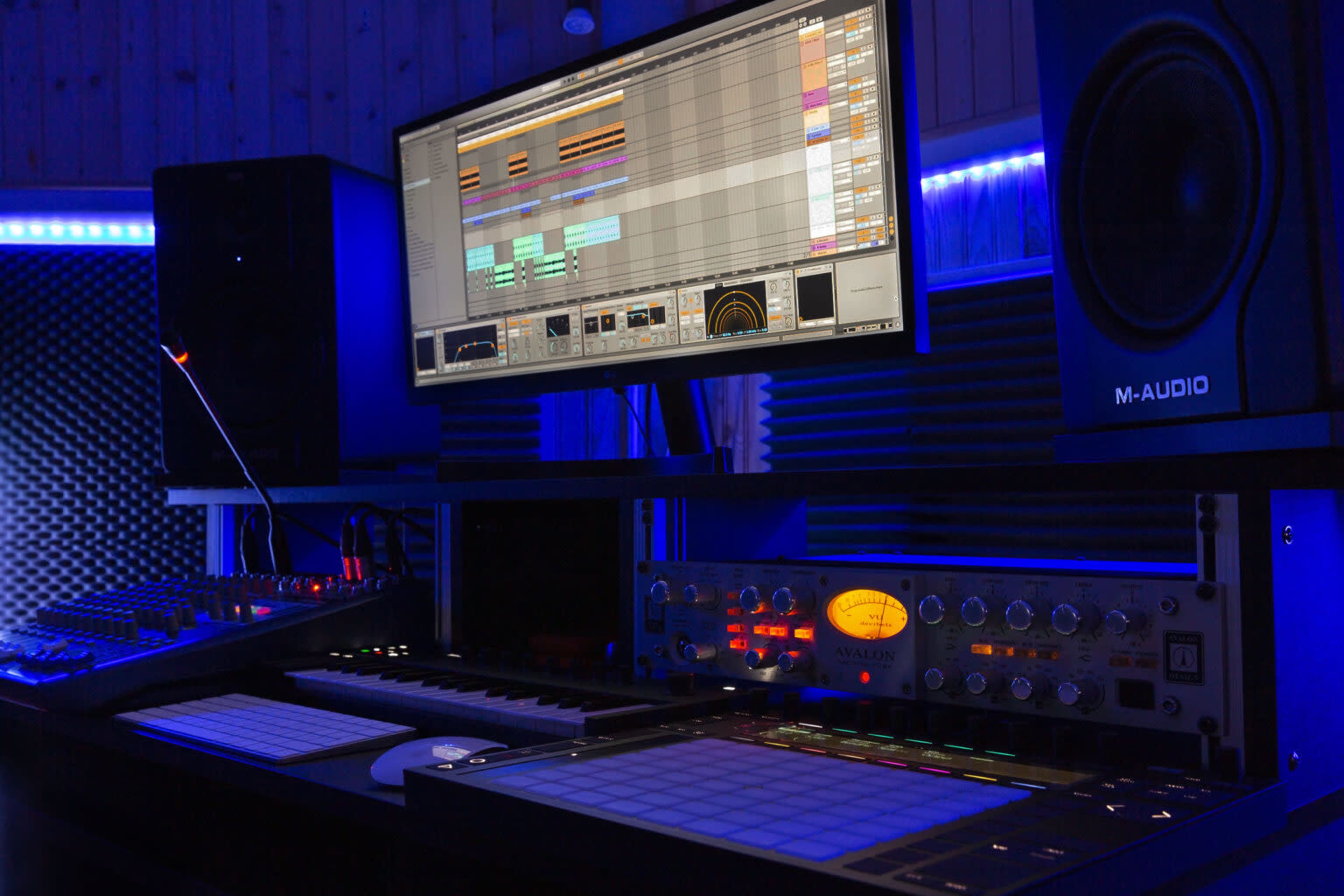 A music production studio with a computer monitor displaying a digital audio workstation, various musical equipment, and ambient blue lighting.