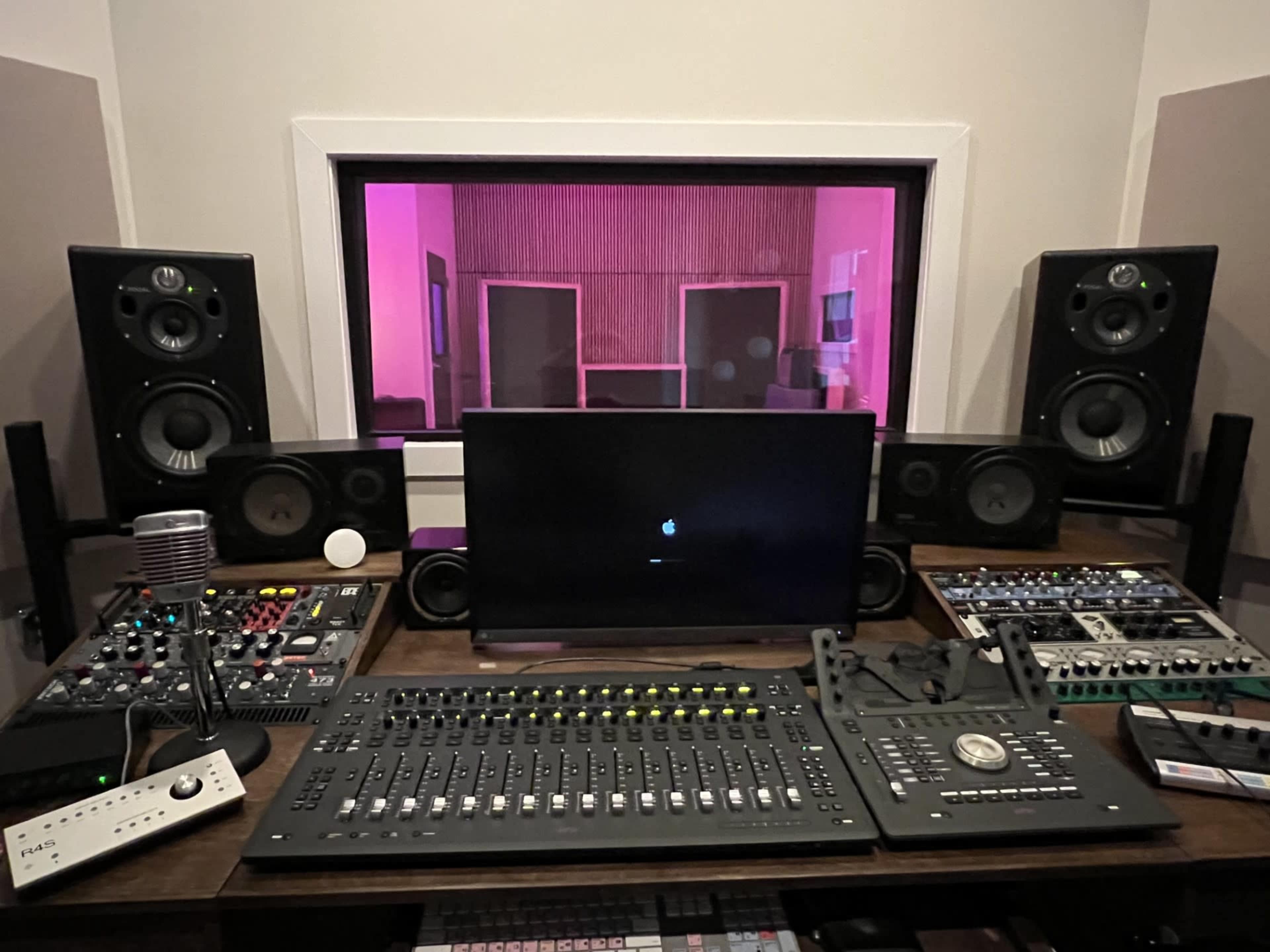 A music production studio with various audio equipment, a digital audio workstation, and a console in front of a glass window.