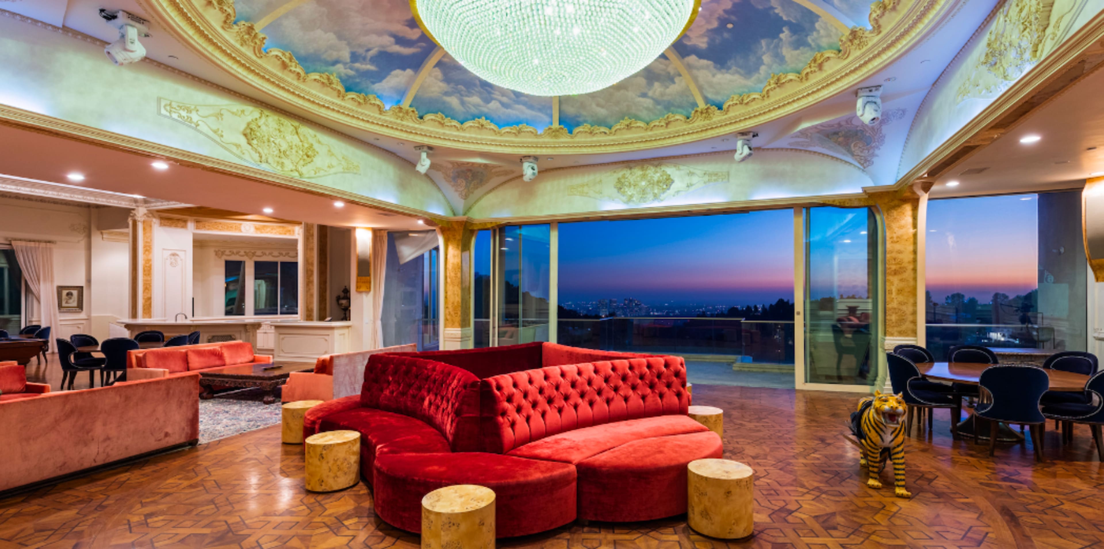 The image shows an elegantly designed lounge with a large chandelier, a vibrant red velvet seating area, and panoramic windows overlooking a sunset view.