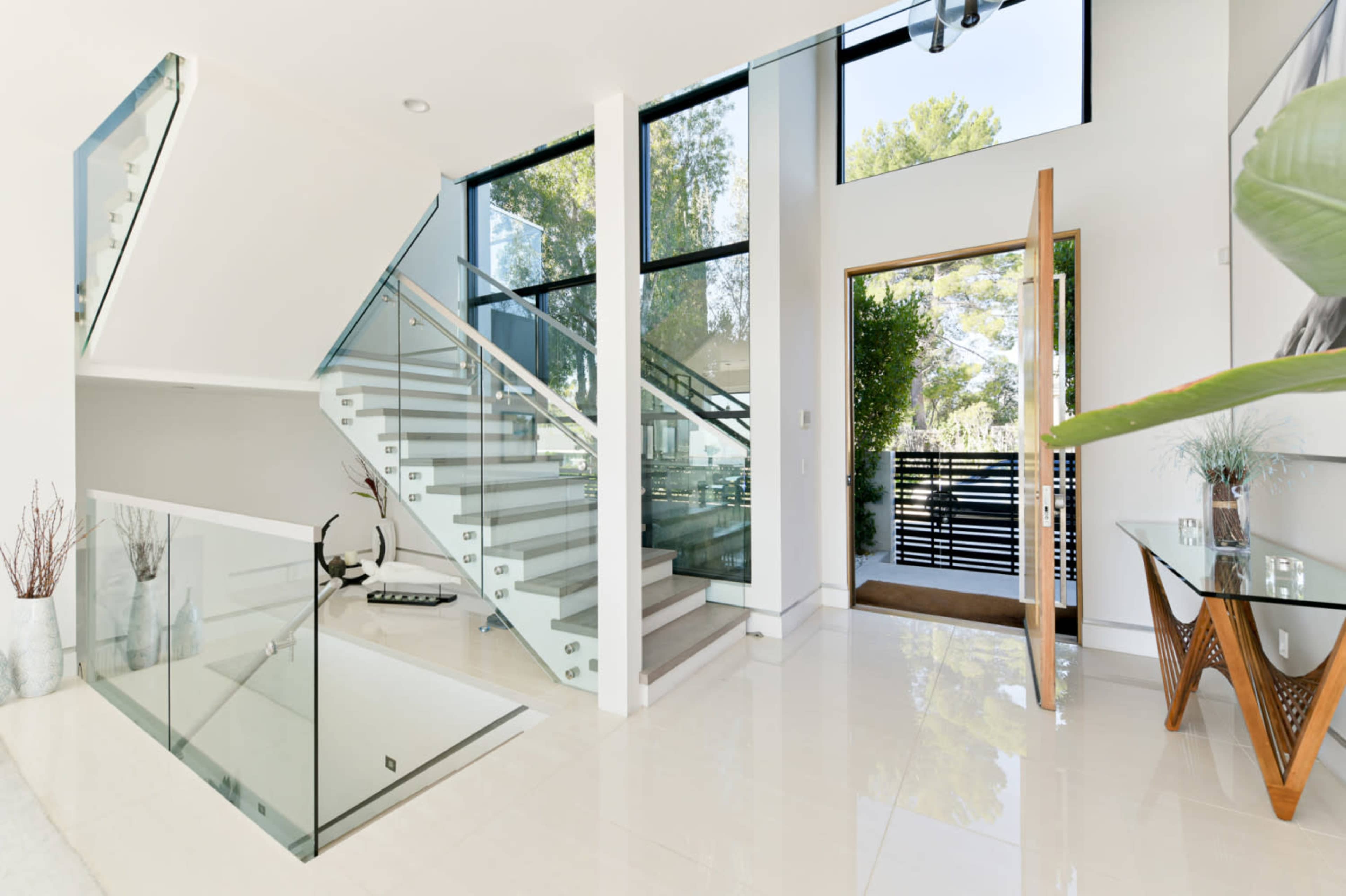 A modern foyer with a glass staircase, large windows, and a wooden front door opening to a bright, spacious entryway.