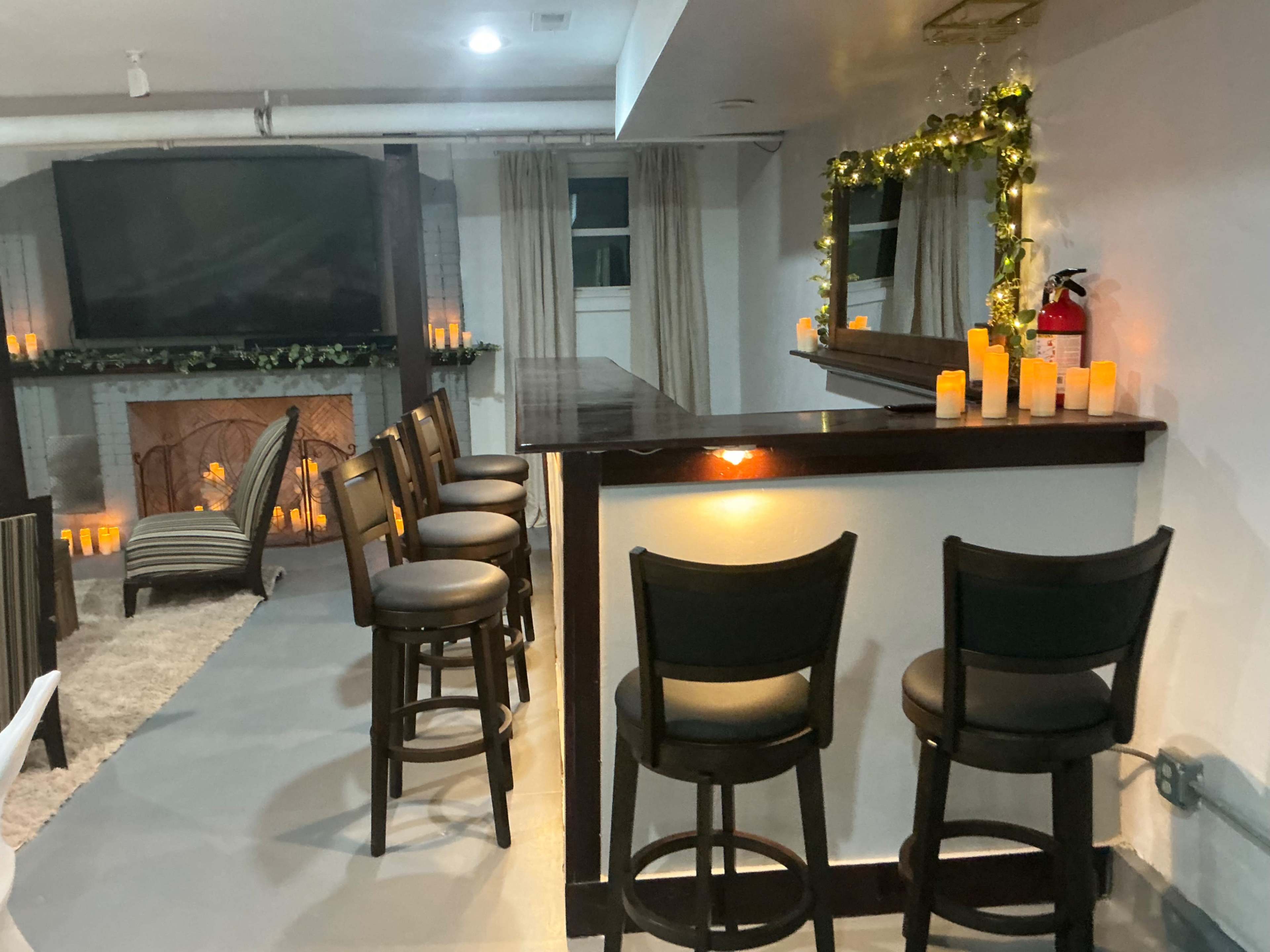 A cozy lounge area features a bar with several stools, a fireplace, and decorative candles, set against a backdrop of a large TV and elegant curtains.