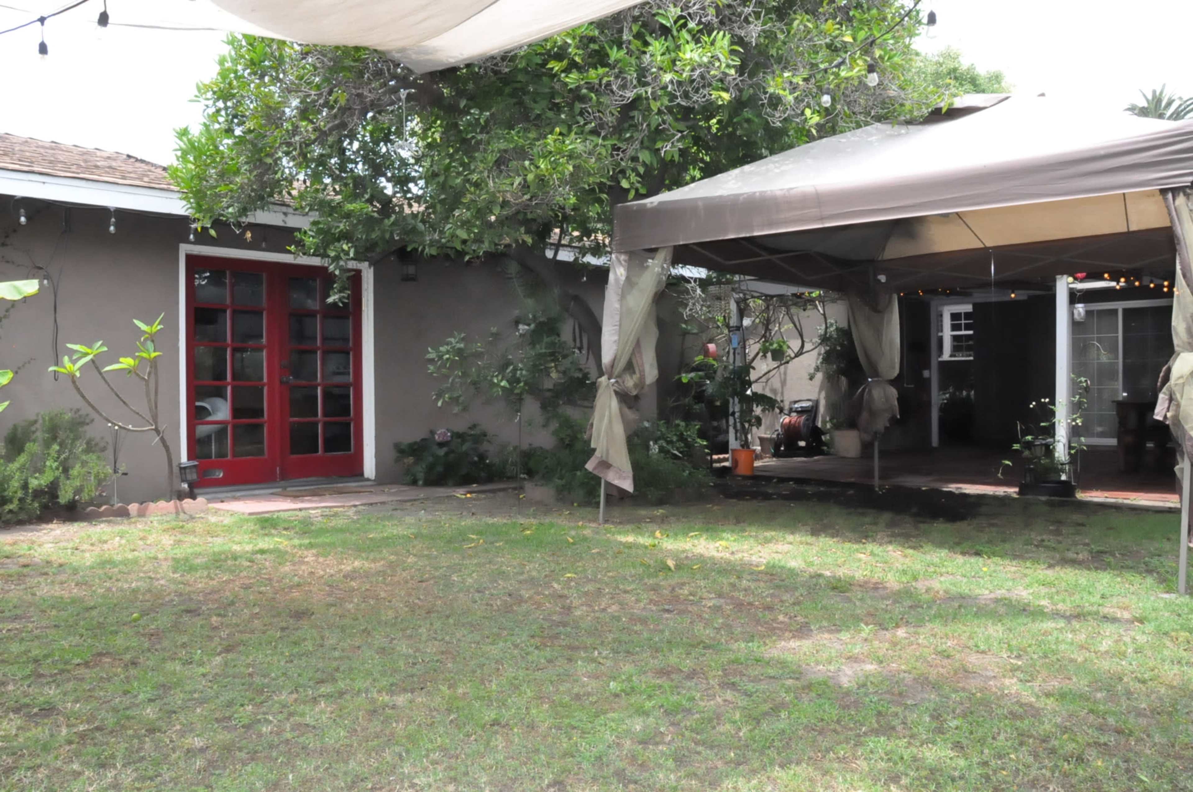 A grassy backyard with a brown house featuring red doors, and a large tent-like structure nearby, surrounded by greenery.